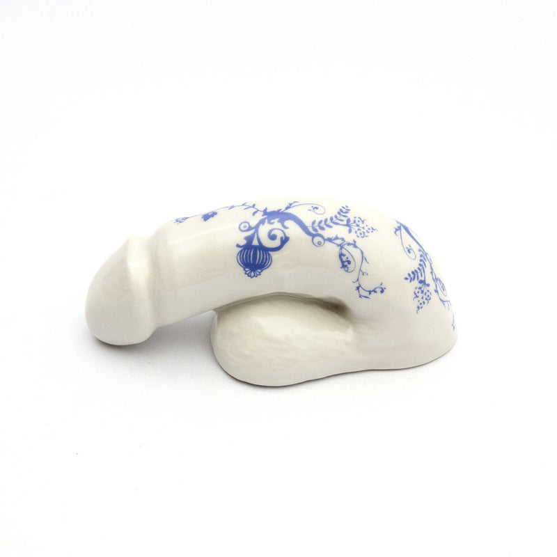 The A Decorative Sex - Blue Onion 3 by Schwanzmanufaktur is a white ceramic sculpture shaped like a banana with intricate blue floral designs, reminiscent of porcelain, and elegantly rests on a smooth white surface.