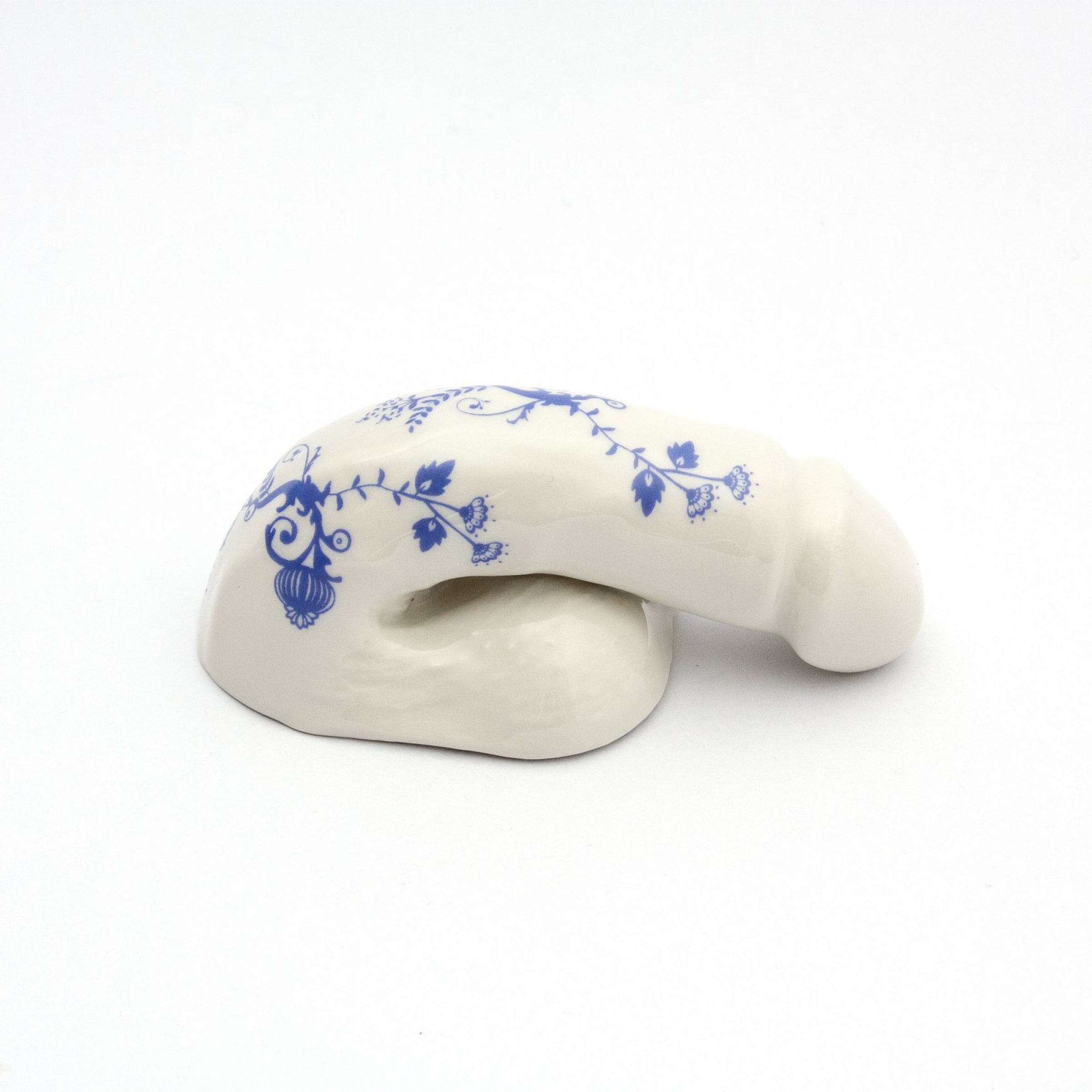 The A Decorative Sex - Blue Onion 3 by Schwanzmanufaktur is a handmade Austrian ceramic sculpture in a flaccid phallic shape, featuring a glossy white finish with intricate blue floral patterns. Its refined elegance and artistry evoke the wit and style of Oscar Wilde.