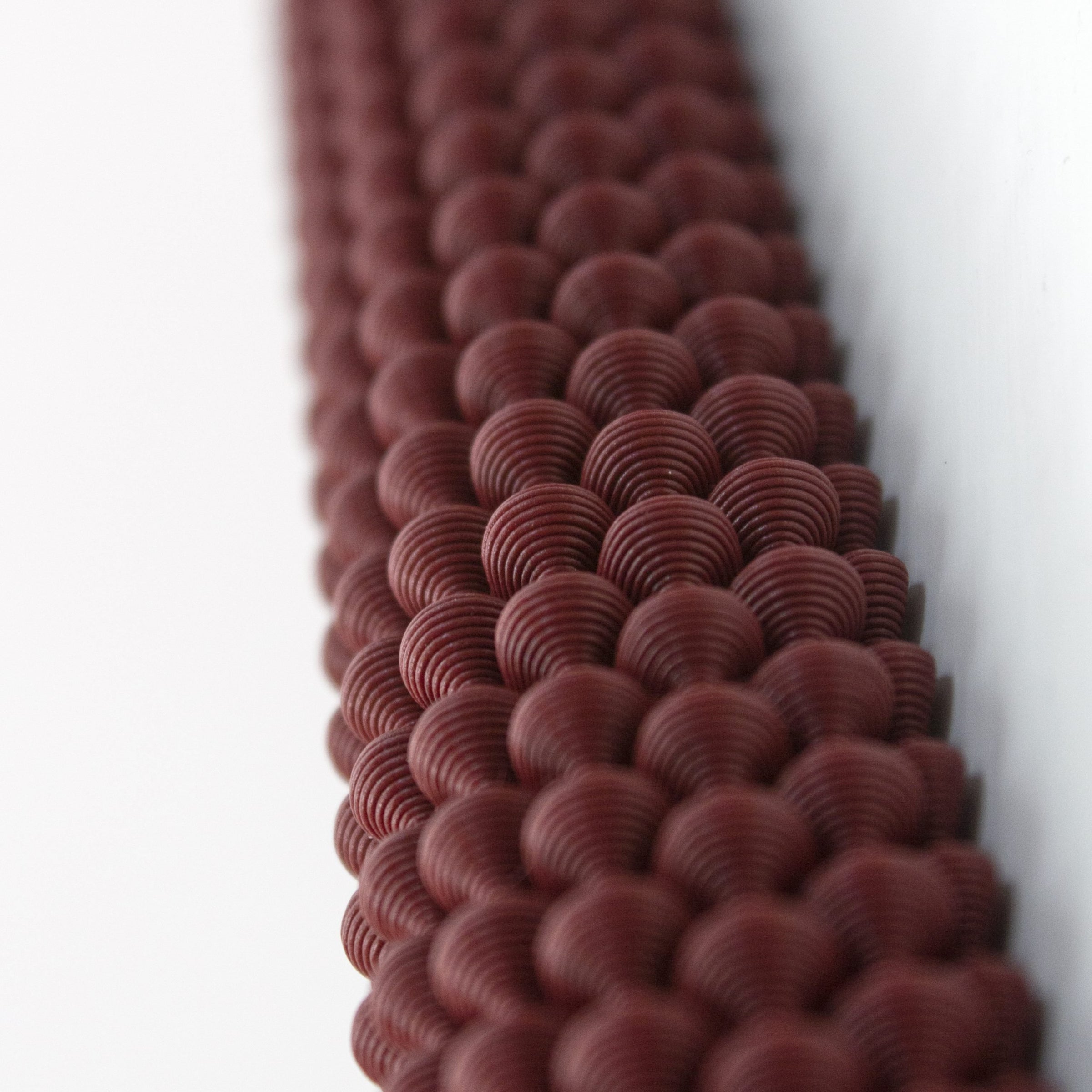 The Hoyt 29g Mezuzah by Sheyn features a close-up of textured dark red spheres, resembling precise 3D printing. The sharp details at the top blur toward the bottom, creating an evocative depth effect.