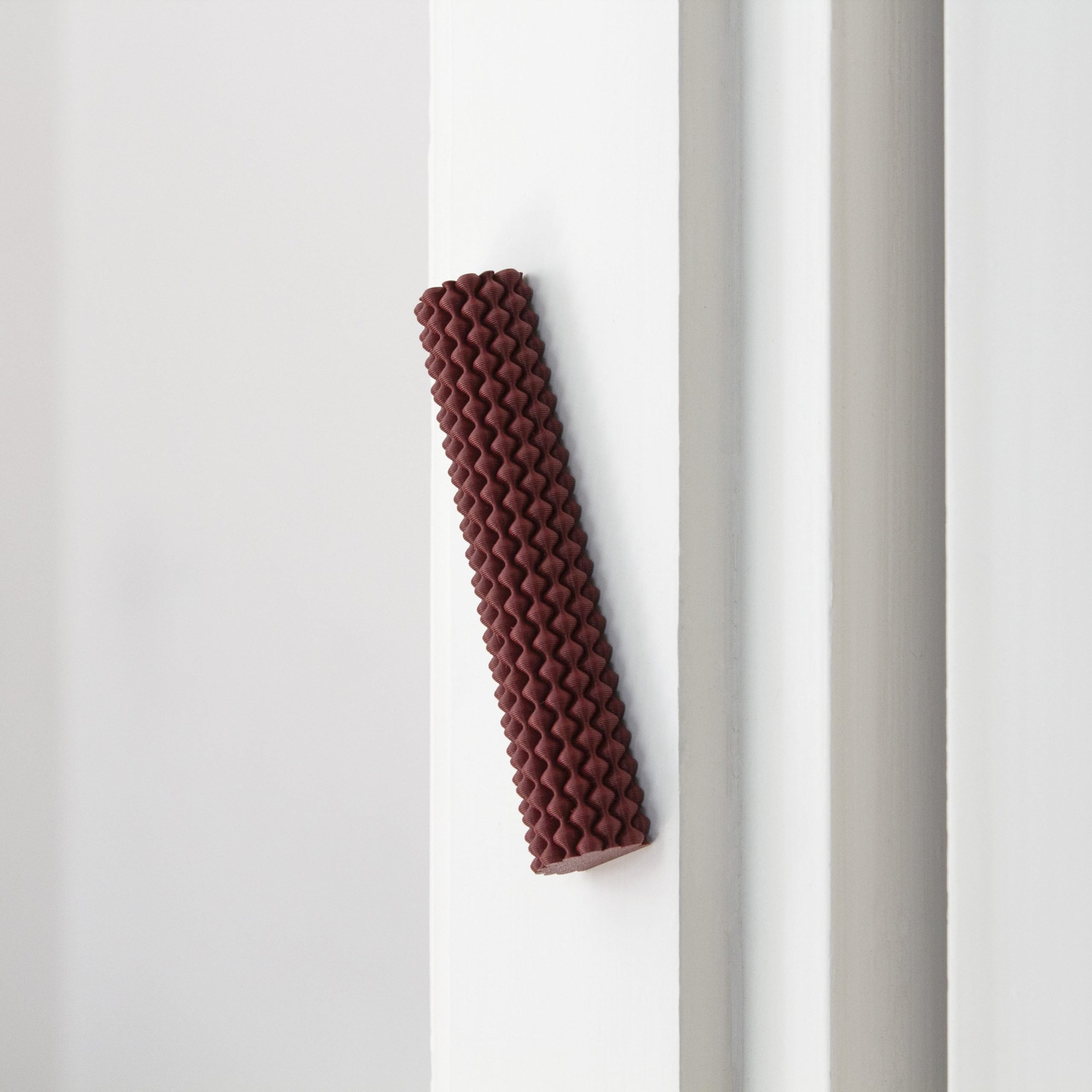 A Hoyt 29g Mezuzah by Sheyn, with its reddish-brown zigzag patterned cylinder, is vertically fixed to a white wall corner. Crafted perhaps through 3D printing in a design studio, it stands out against the walls smooth surface.