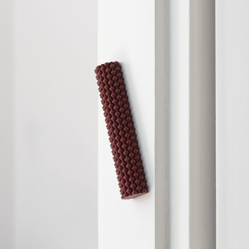 A Hoyt 29g Mezuzah by Sheyn, with its reddish-brown zigzag patterned cylinder, is vertically fixed to a white wall corner. Crafted perhaps through 3D printing in a design studio, it stands out against the walls smooth surface.