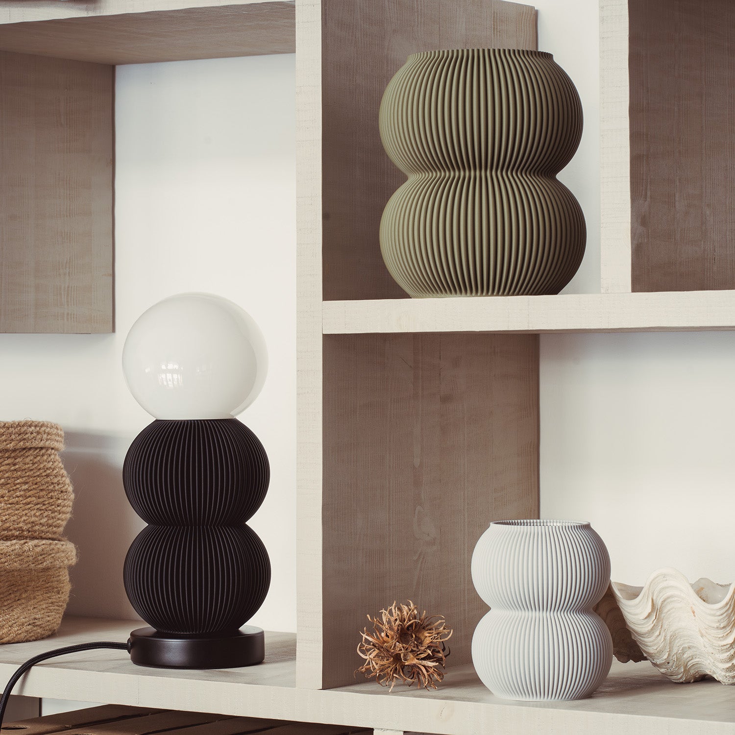 A black lamp with a round white bulb rests on a wooden shelf. Next to it, dried decorative foliage accompanies an eco-friendly Zayl 159g Vase by Sheyn. On the upper shelves are a green ribbed vase and a large seashell next to a woven basket made from recyclable materials.