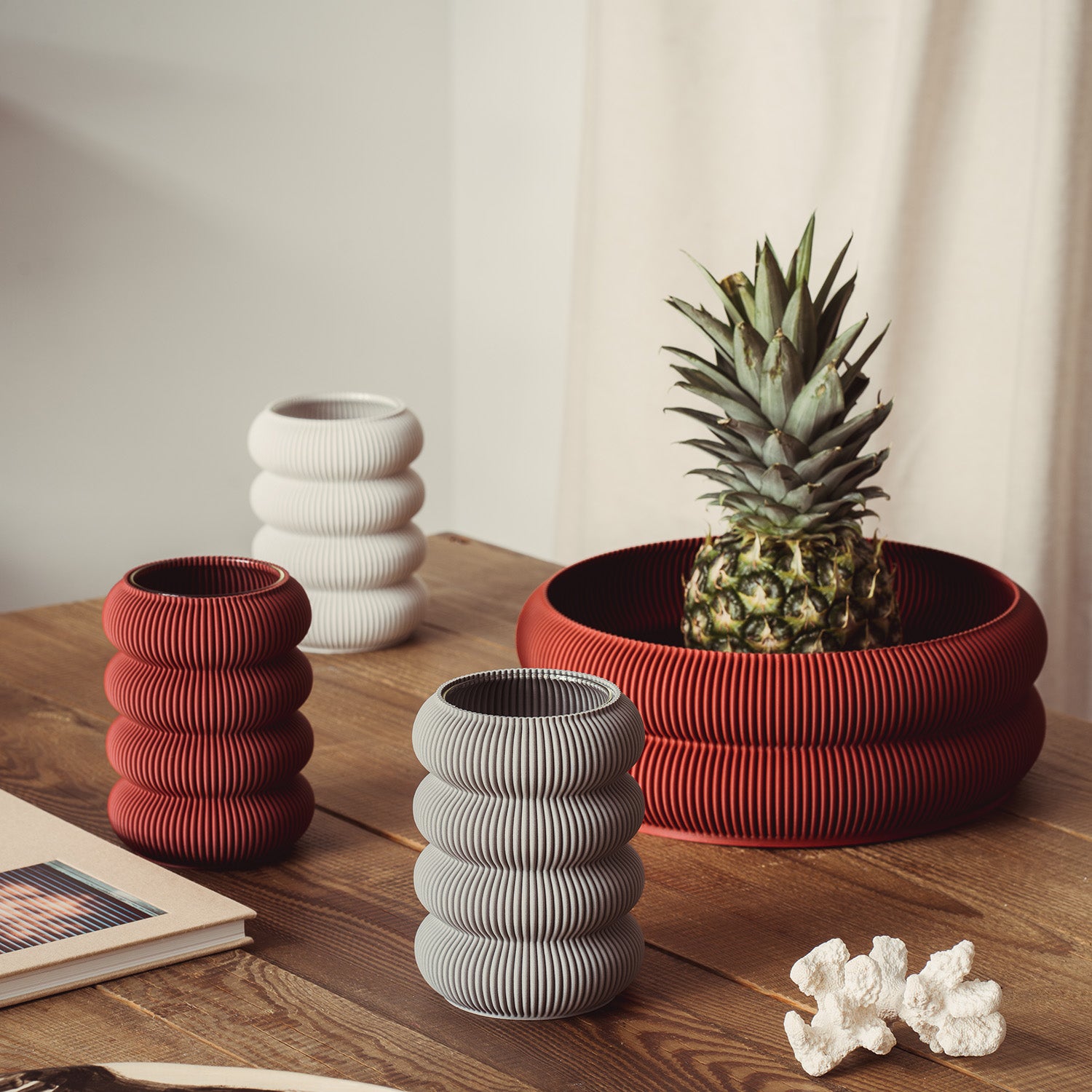 Sheyns Zayl 130g vases, with their ribbed textures reminiscent of architectural pillars, in red, white, and gray are arranged on a wooden table. A red ribbed bowl holds a pineapple alongside a book and decorative coral piece, set against a cream curtain backdrop.