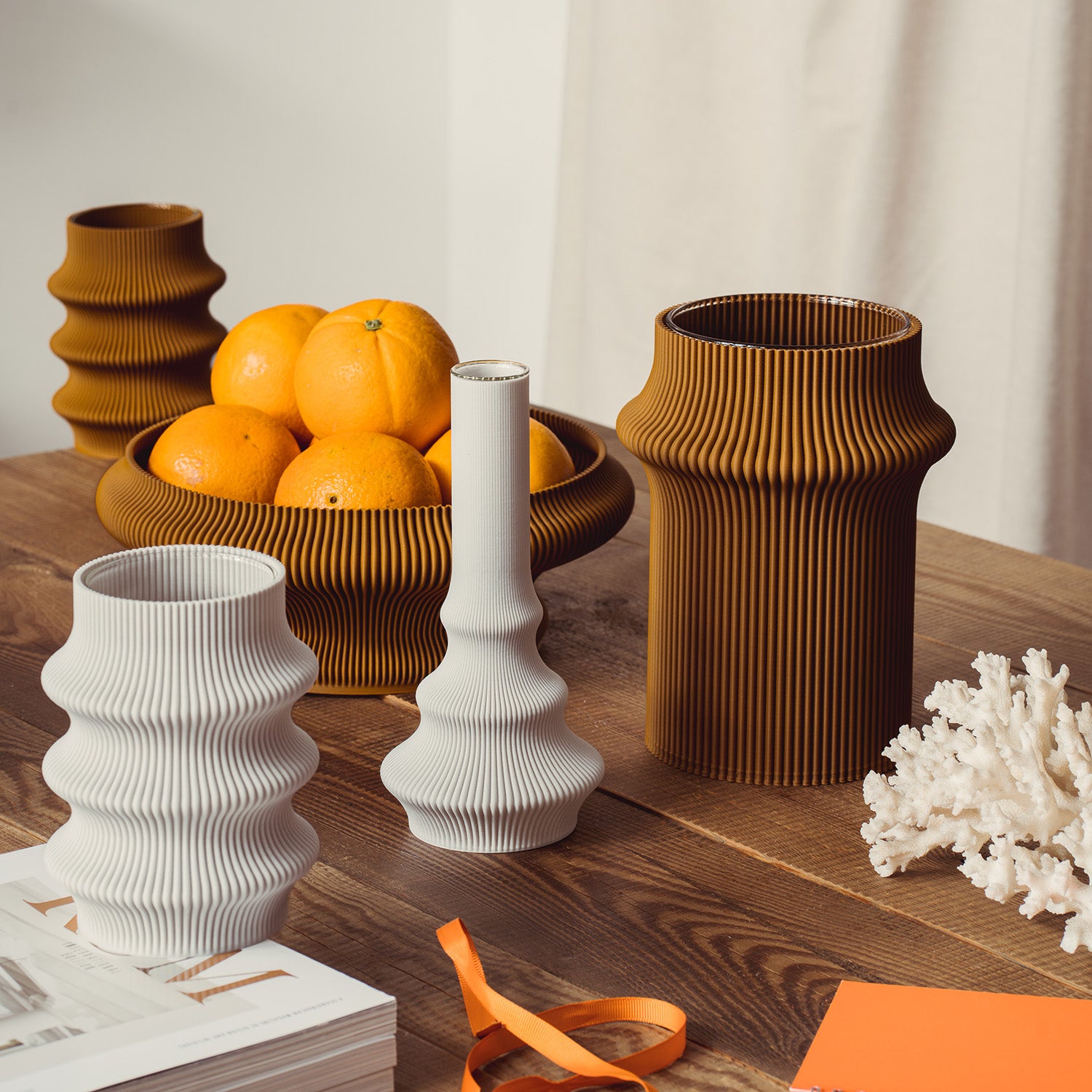 A wooden table displays three Zayl 247g vases by Sheyn, resembling architectural pillars in white and brown. An eco-friendly bowl holds oranges, with a white coral decor and books nearby. The minimalistic setting is enhanced by soft, natural lighting.