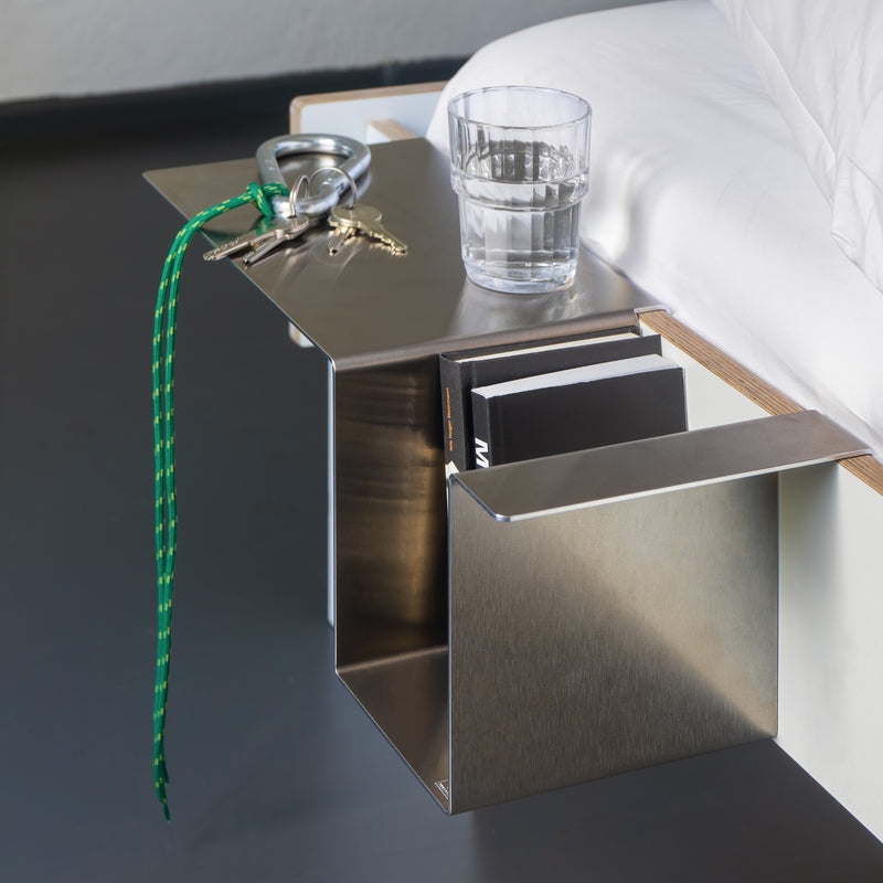 The Moormann Siebenschläfer, a minimalist metal bedside shelf, easily attaches to a flat-packed bed without tools. It holds essentials like a glass of water, keys with a green lanyard, and two books. The shelf elegantly extends over the side against the contrast of a dark floor and white wall.