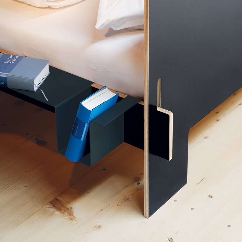 The Siebenschläfer by Moormann is a sleek black-framed, modern flat-packed bed on a birch plywood floor. It includes a built-in shelf at the foot holding three colorful books. The partially visible mattress and white bedding require no tools for assembly.