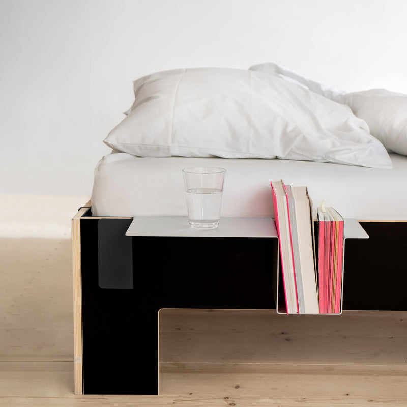 The minimalist bedroom features Moormanns Siebenschläfer, a modern, tool-free, flat-packed platform bed crafted from birch plywood. A black-and-white side table attached to the bed holds a glass of water and pink and white books. Neutral-toned bedding rests on the wooden floor.