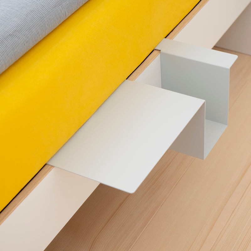 The Siebenschläfer by Moormann features a modern bed corner with a bright yellow mattress and an L-shaped white birch plywood shelf. Its flat-packed design highlights simple, functional elegance and sits on a light hardwood floor, all assembled without any tools.