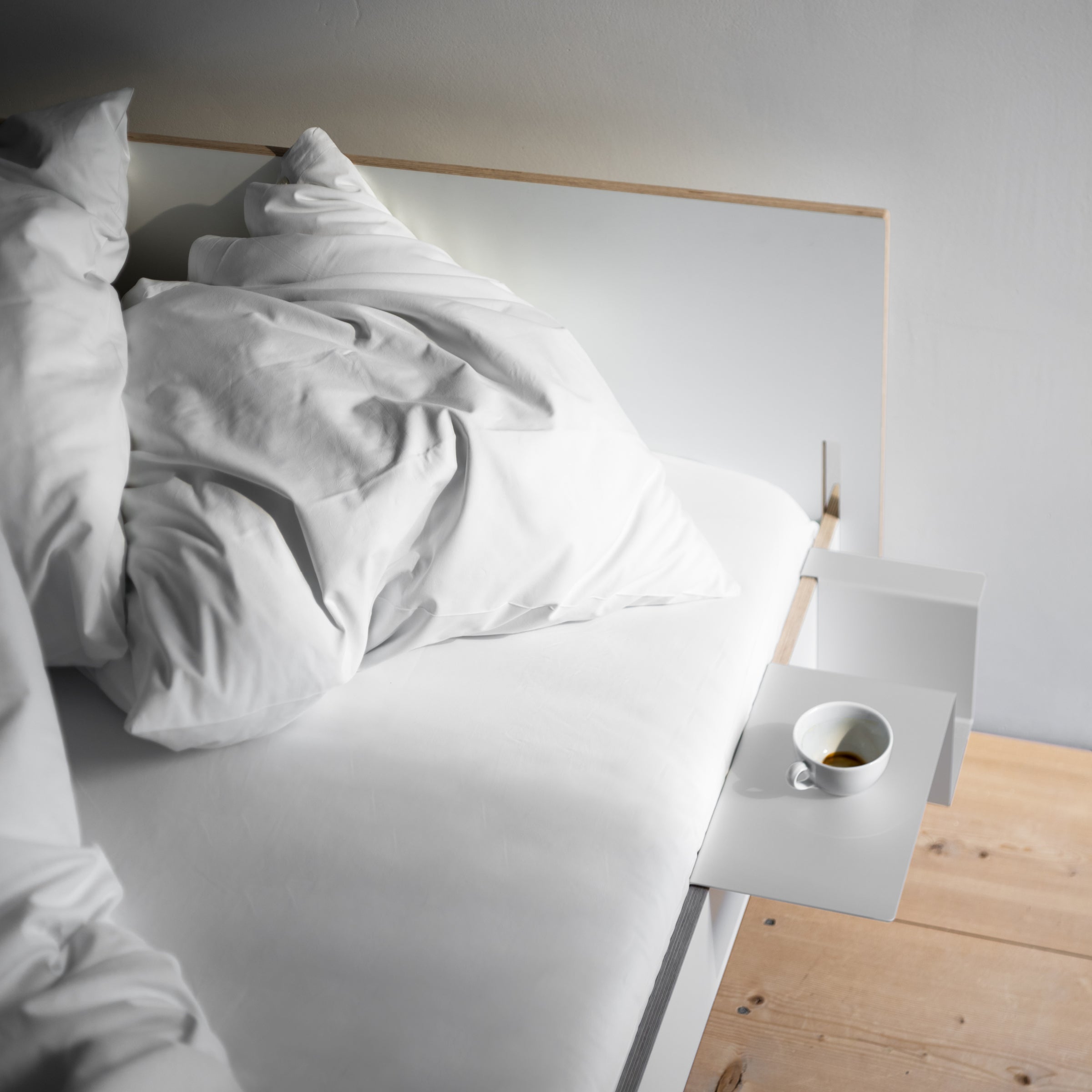 The Siebenschläfer by Moormann is a neatly crafted flat-packed bed, featuring white pillows and sheets. Attached is a modern white side table with an espresso cup. The room includes birch plywood wooden flooring and a light grey wall.