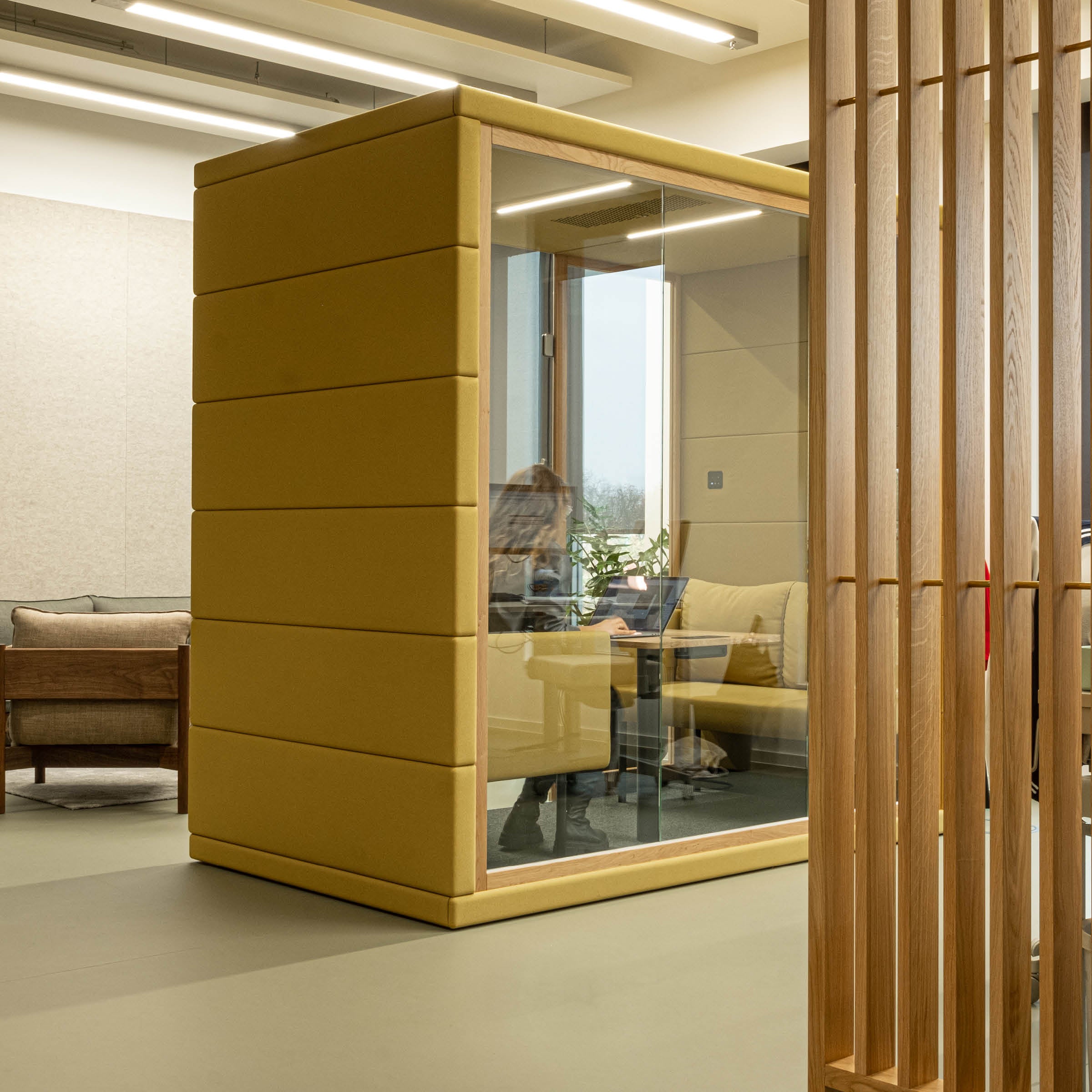 In the compact, yellow OFFICE POD QUADRIO 1 by SilentLab, a person enjoys privacy at a desk and chair. The soundproof booth with glass doors is set in a modern office, surrounded by sleek wood paneling and comfy seating.