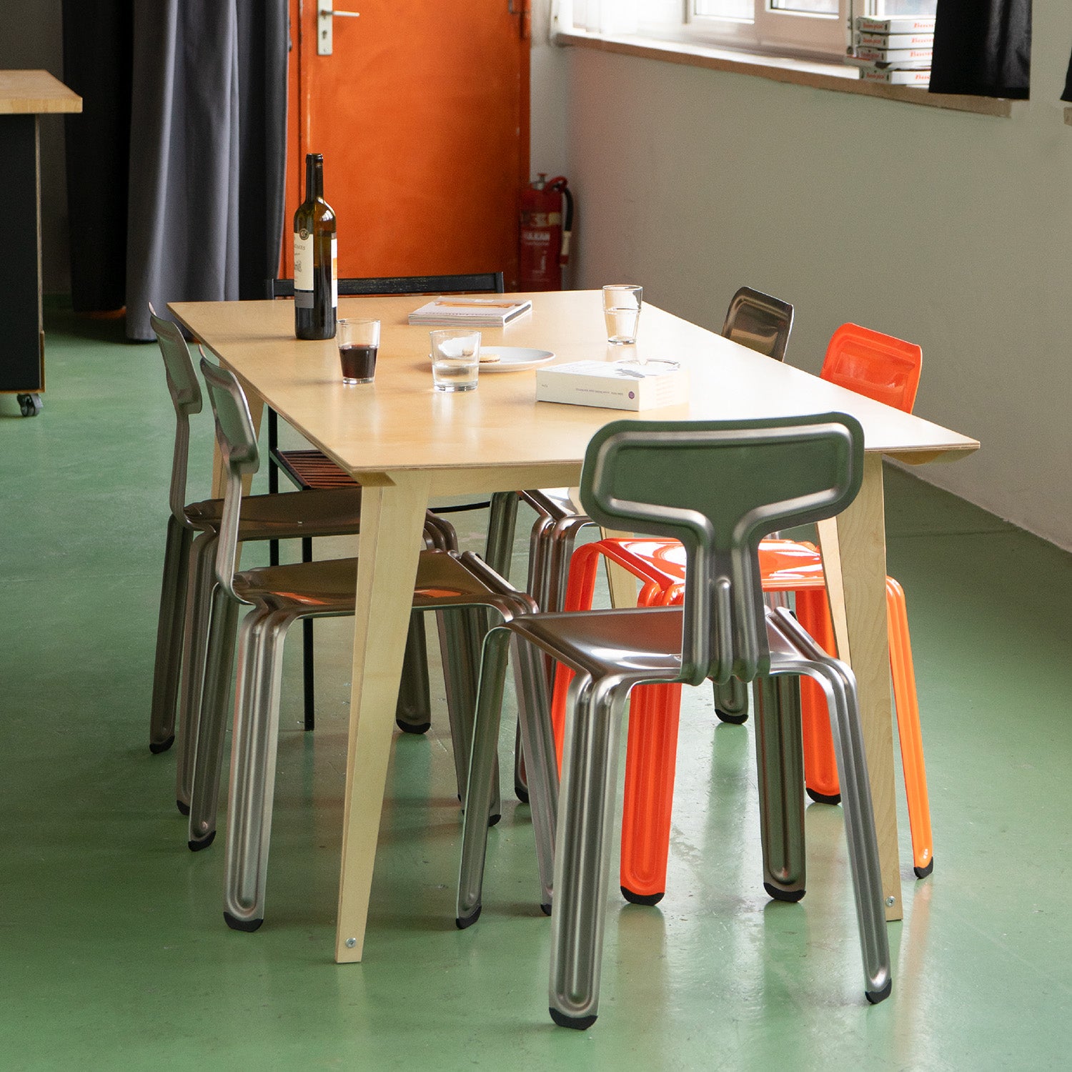 The Moormann Spanoto wooden dining table made from birch plywood is paired with four metallic chairs—two orange, two silver. It stands on a green floor, holding a wine bottle, water glasses, and a book. Sunlight streams through a window near the closed orange door in the background.