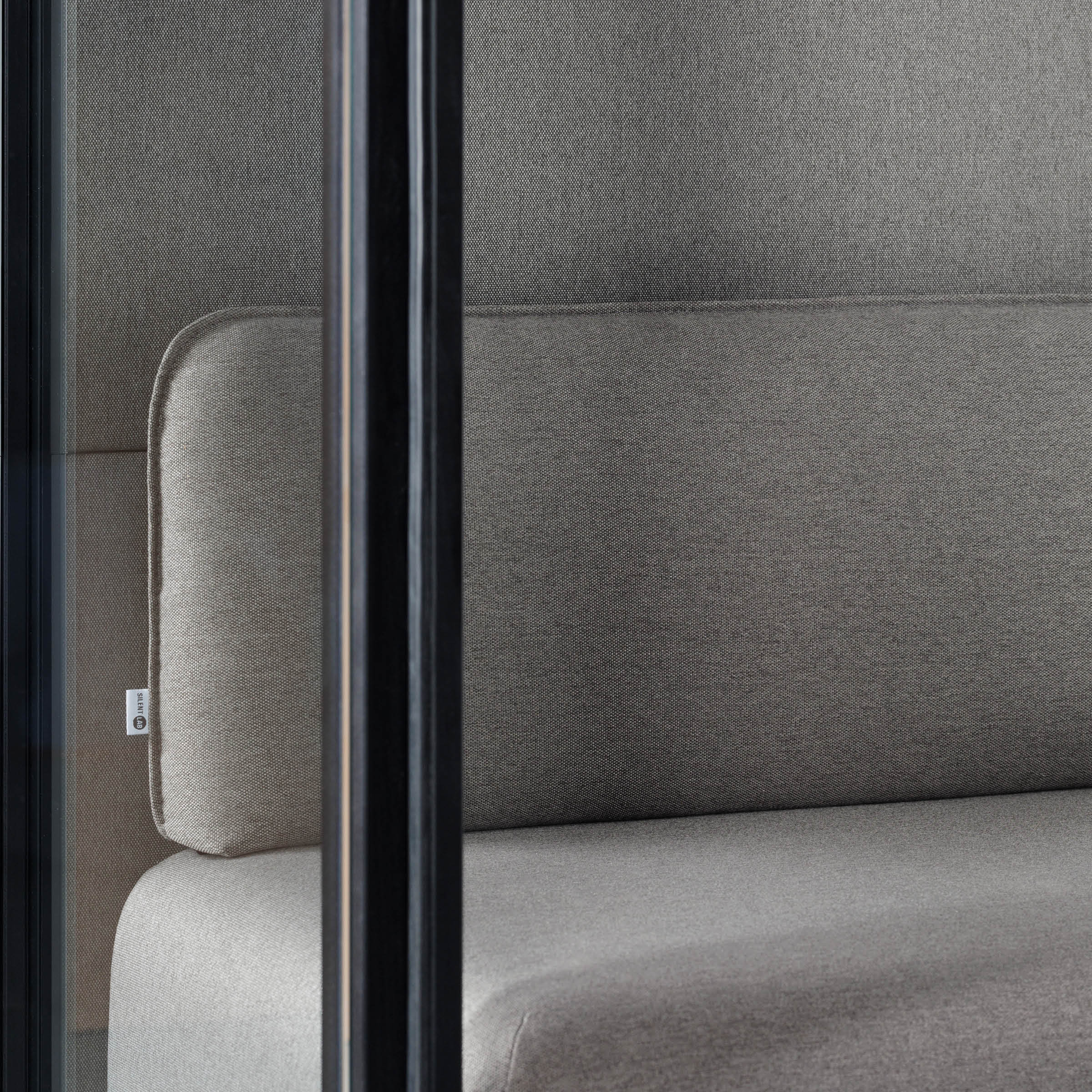 The OFFICE POD SPARK 2 by SilentLab features a modern, gray upholstered couch within its sleek design. Enclosed by glass panels, the pods neutral colors and texture offer a minimalist aesthetic while enhancing acoustic privacy for better working conditions.