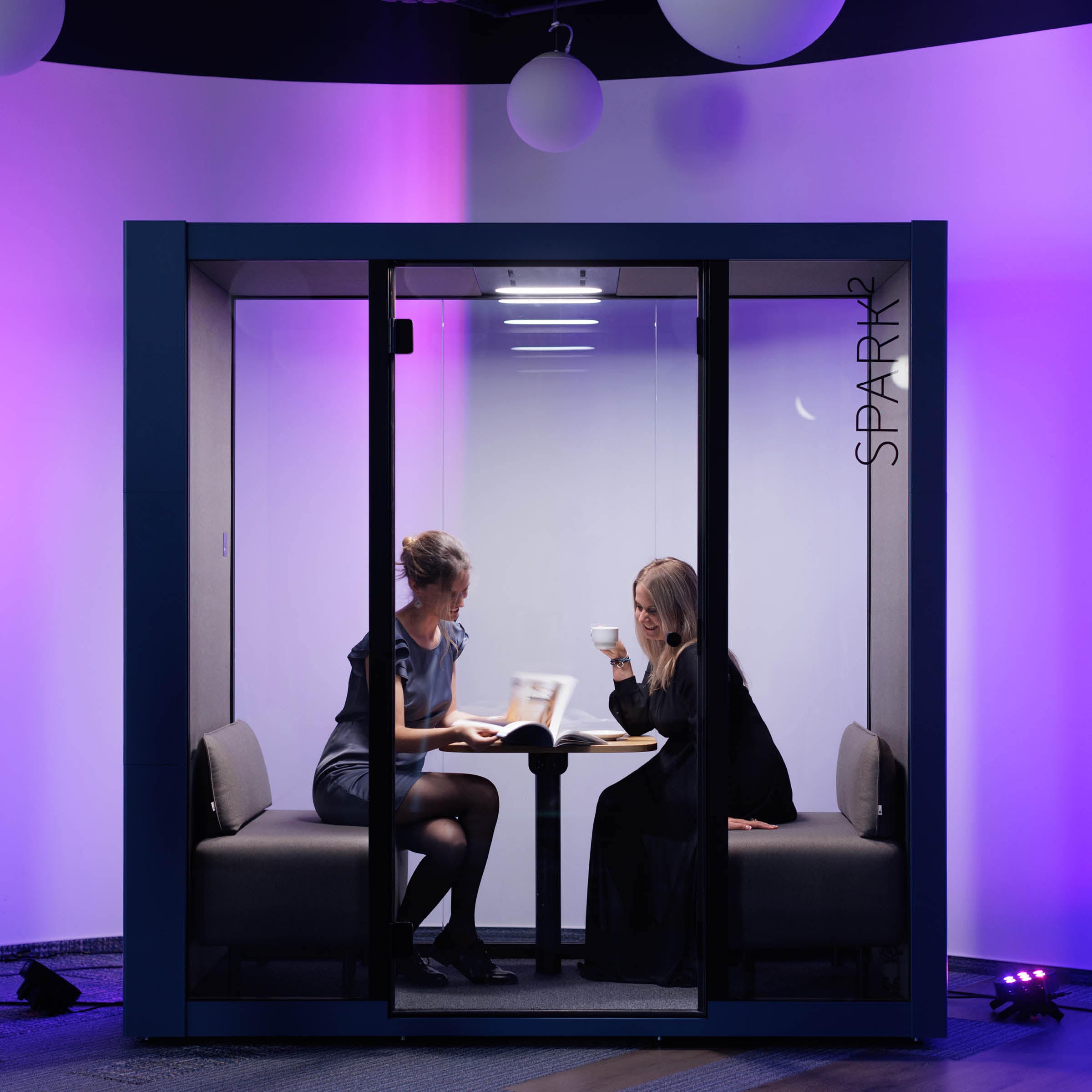 Two people sit inside the SilentLab OFFICE POD SPARK 2 with glass walls, reading and sipping coffee. The pods soothing purple lighting creates a serene atmosphere against the dark background, enhancing their working conditions.