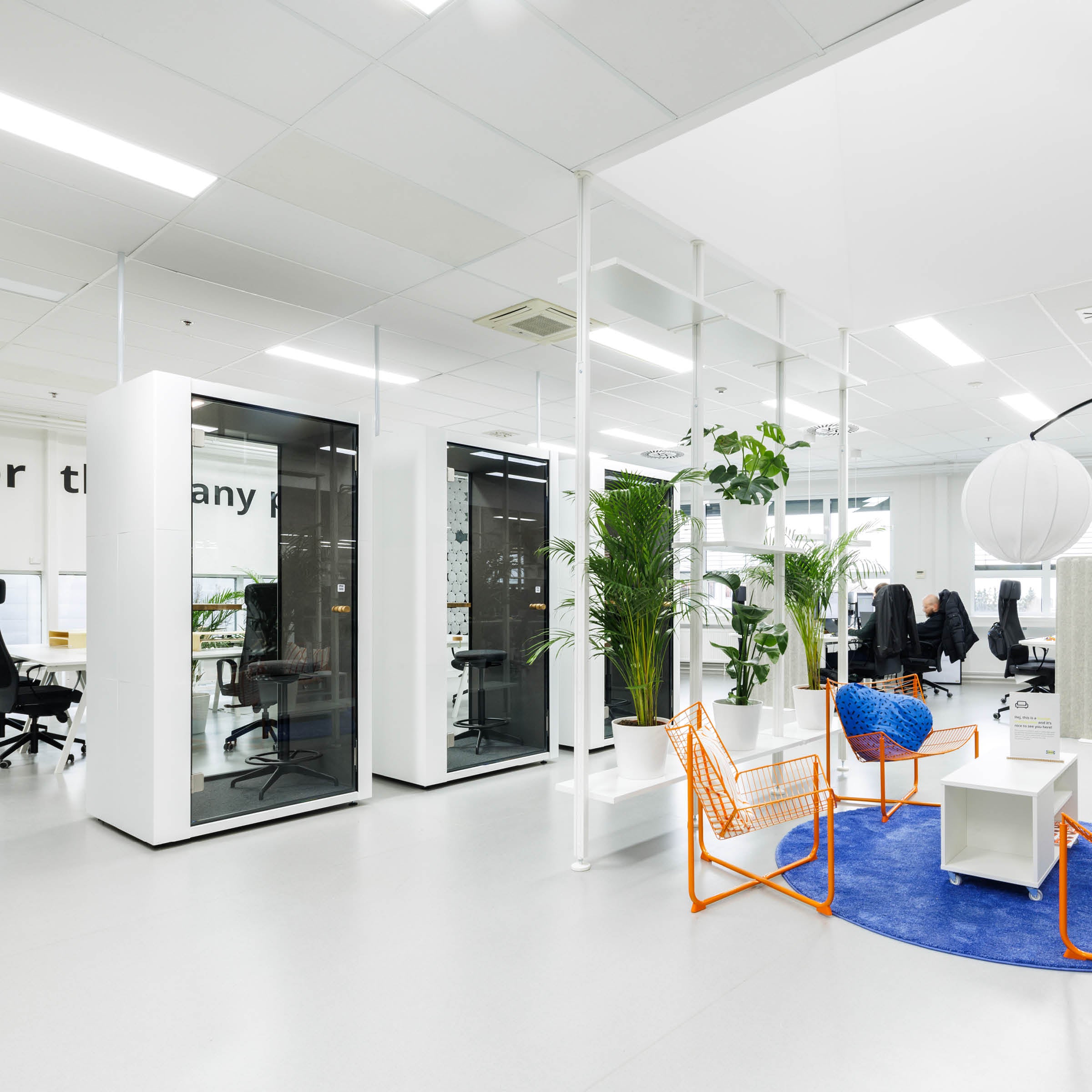 Modern office space featuring the SilentLab OFFICE POD SPARK, desks, and chairs in a bright, open area. Potted plants decorate the room, while a round blue rug with orange chairs adds color. Large white lanterns hang from the ceiling, enhancing the rooms contemporary vibe.