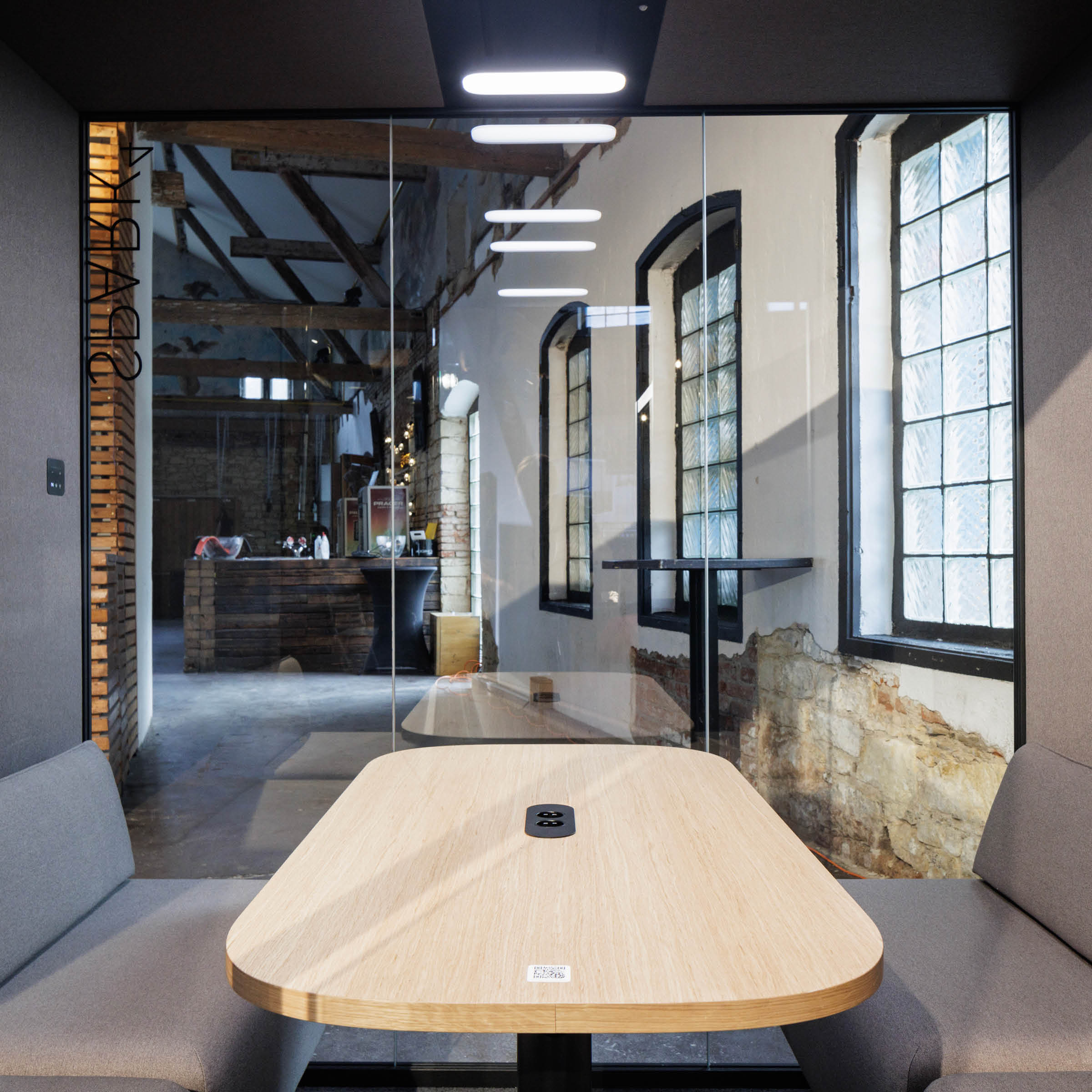 A modern office features a small meeting room with a wooden table and gray cushioned benches. A glass partition overlooks a room with large windows, exposed brick, and wooden beams. Equipped with SilentLabs OFFICE POD SPARK 4 for private meetings, ceiling lights ensure brightness.