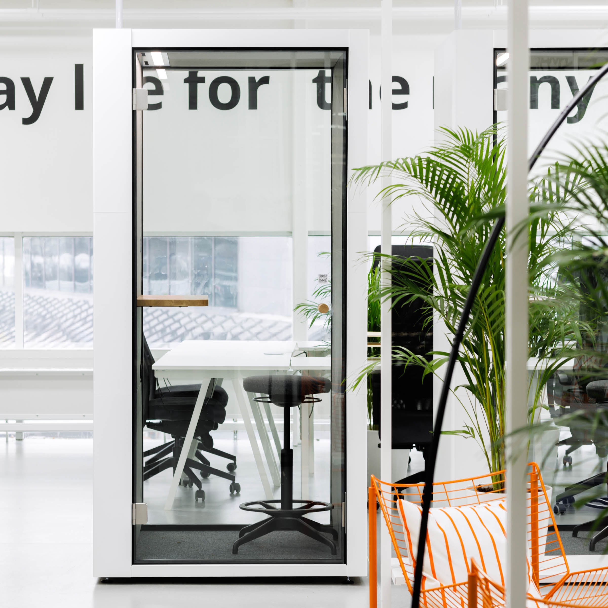 A modern office features SilentLabs OFFICE POD SPARK, a glass office pod with a wooden stool and table. Nearby, office chairs, potted plants, and large windows enhance the ambiance. A wall showcases bold black text for added flair.