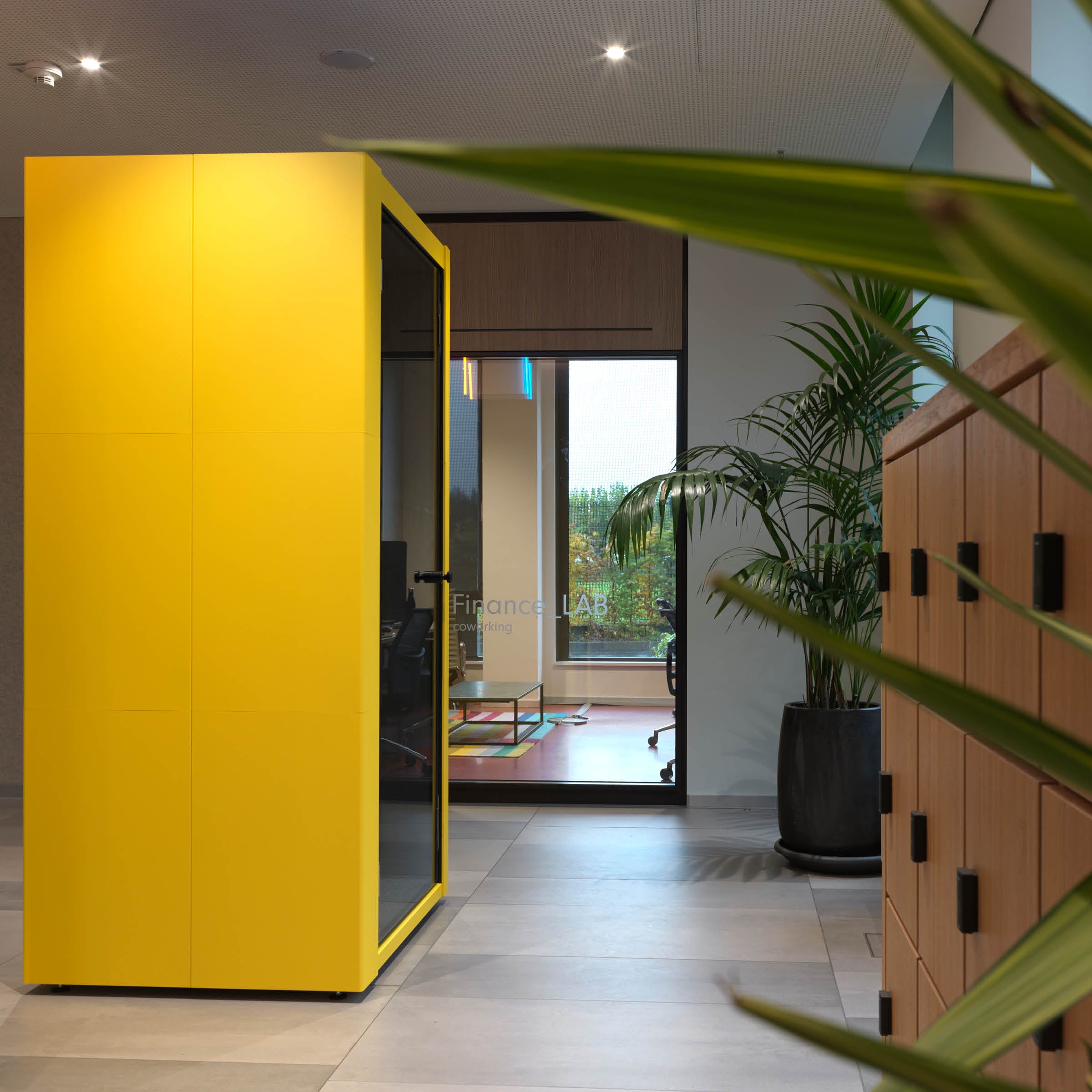 The modern office space showcases SilentLabs large, yellow OFFICE POD SPARK with wooden lockers on the right. A glass door in the background leads to a conference room with visible chairs and a plant. The area is spacious and well-lit.