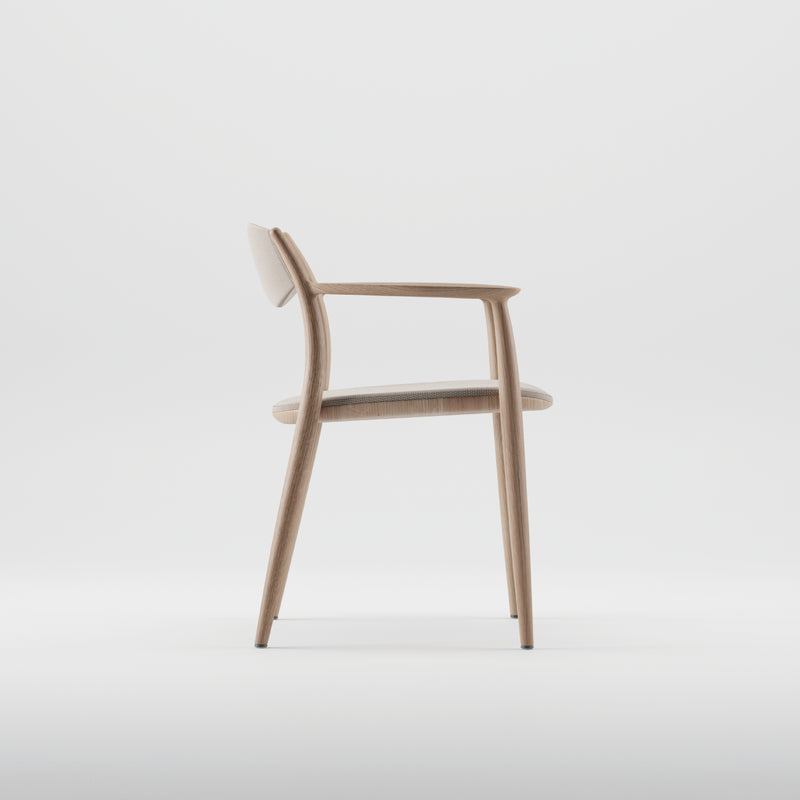 The NILA Chair by Artisan features a side view of a minimalist design, crafted from solid wood and finished with natural oils. It has a light wooden frame, upholstered backrest, and seat cushion. Slim, angled legs enhance its clean aesthetic using environmentally-friendly materials.
