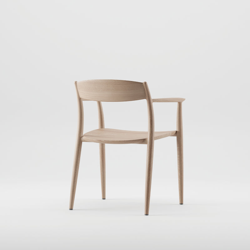 The NILA Chair by Artisan is a minimalist wooden piece with a light finish, crafted from solid wood and enhanced with natural oils. It features a curved backrest, slender legs, and simple armrests against a plain white background.