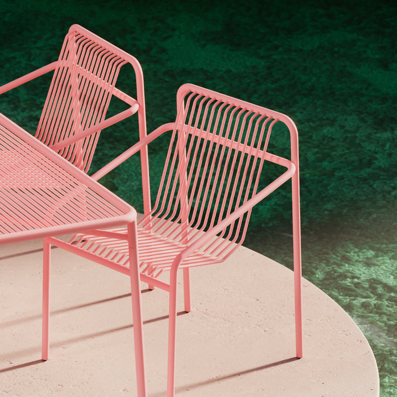The IVY Outdoor Dining Chair by Objekte unserer Tage shows off its pink wireframe design alongside a matching angular table. The set is weather-resistant, elegantly set on a light circular surface, contrasting with the dark green background for a blend of style and durability.