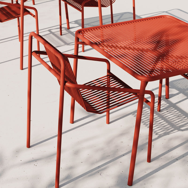 The IVY Outdoor Dining Chair set from Objekte unserer Tage, featuring red metal chairs and a square table, sits on a light surface with shadows cast in sunlight. The minimalist linear design is crafted from weather-resistant materials for lasting style.