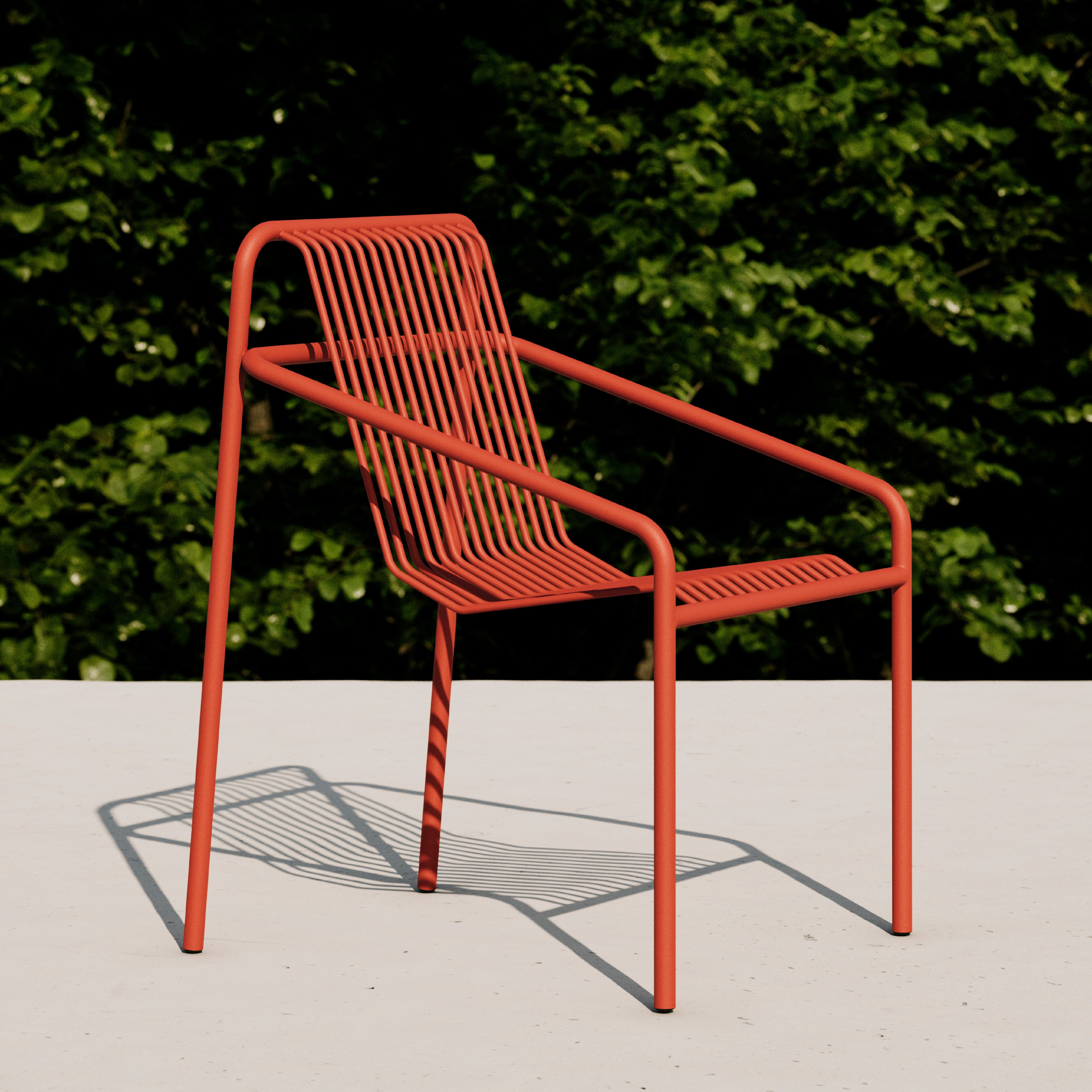 The Objekte unserer Tage IVY Outdoor Dining Chair, a modern red metal piece with a minimalist slatted design, stands on a light surface. The sun casts intricate shadows below as blurred green foliage in the background highlights its weather-resistant materials.