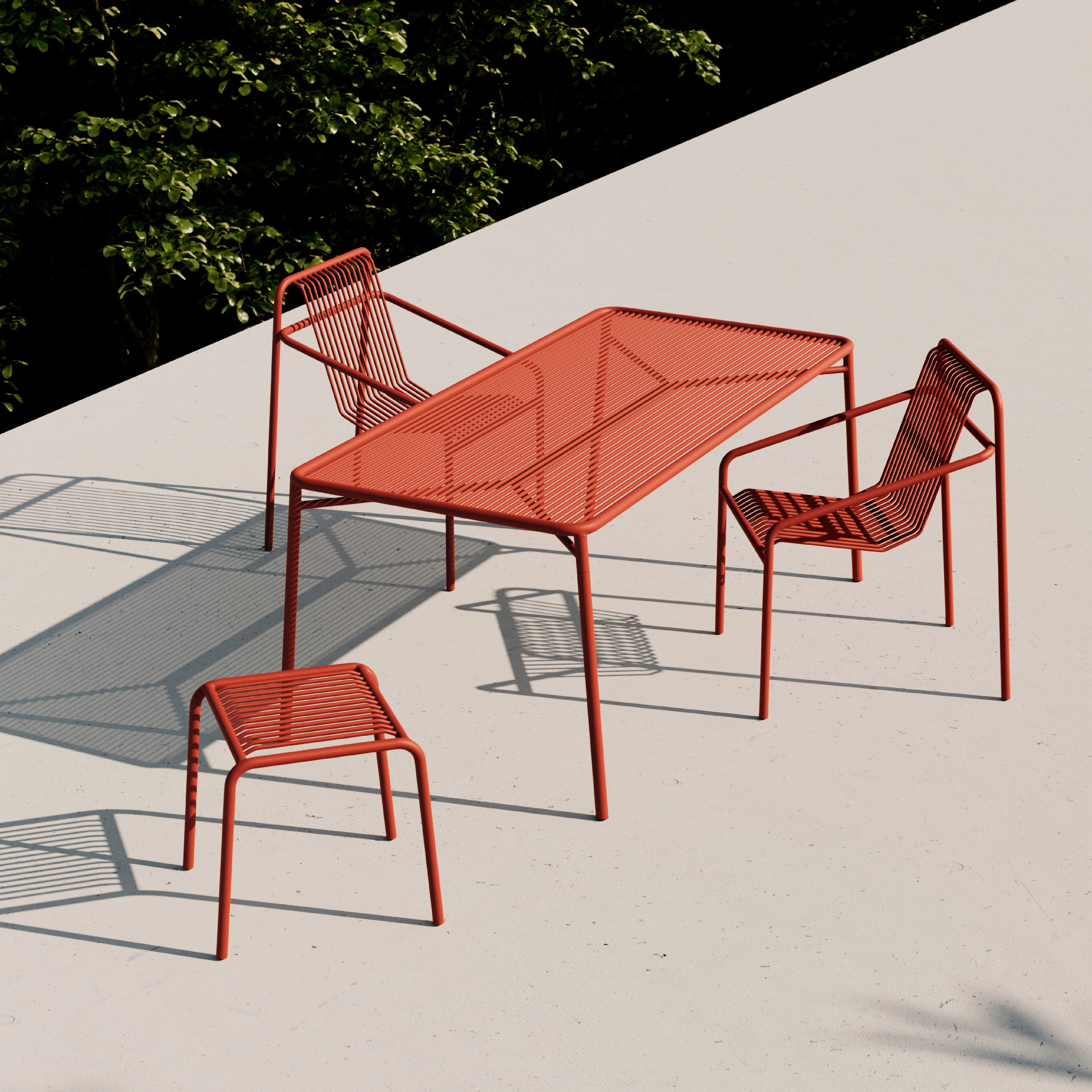 The IVY Outdoor Stool by Objekte unserer Tage complements a rooftop setup with a red powder-coated steel table and two chairs. The wireframe design creates intricate shadows, while green foliage accents the background.