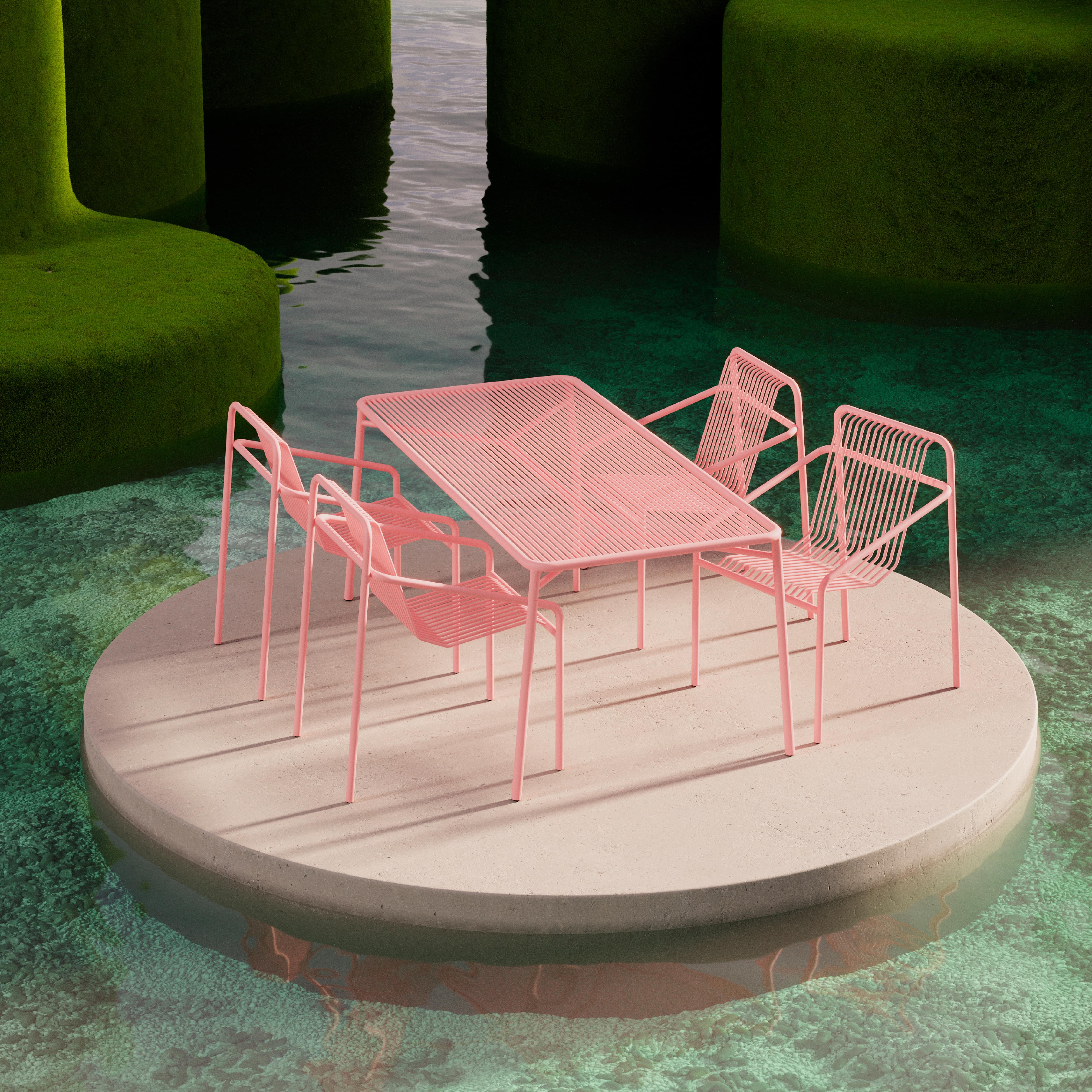 The IVY Outdoor Table by Objekte unserer Tage, in stylish pink metal, features four stackable chairs set on a circular concrete platform surrounded by water. Its powder-coated steel ensures resilience, while lush green formations rising from the water create a surreal and tranquil setting.