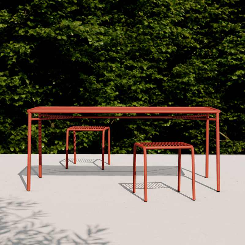 The IVY Outdoor Table by Objekte unserer Tage, crafted from rust-colored powder-coated steel, features a minimalist design alongside two stackable stools. They complement a white patio beautifully with lush green foliage providing a serene backdrop.