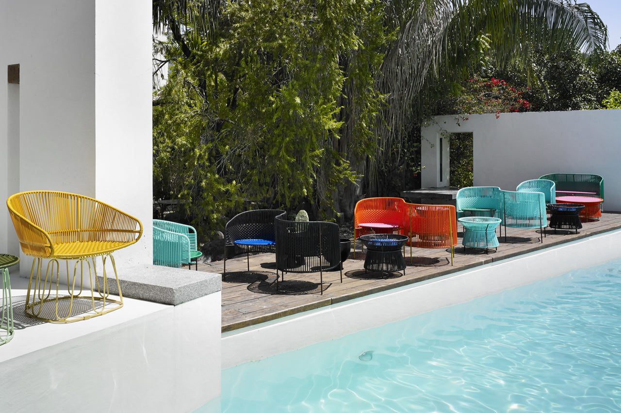 A poolside scene with various colorful chairs and tables arranged on a wooden deck. The pools clear blue water is visible in the foreground, surrounded by lush greenery. A single yellow chair is placed near the pools edge on the left.