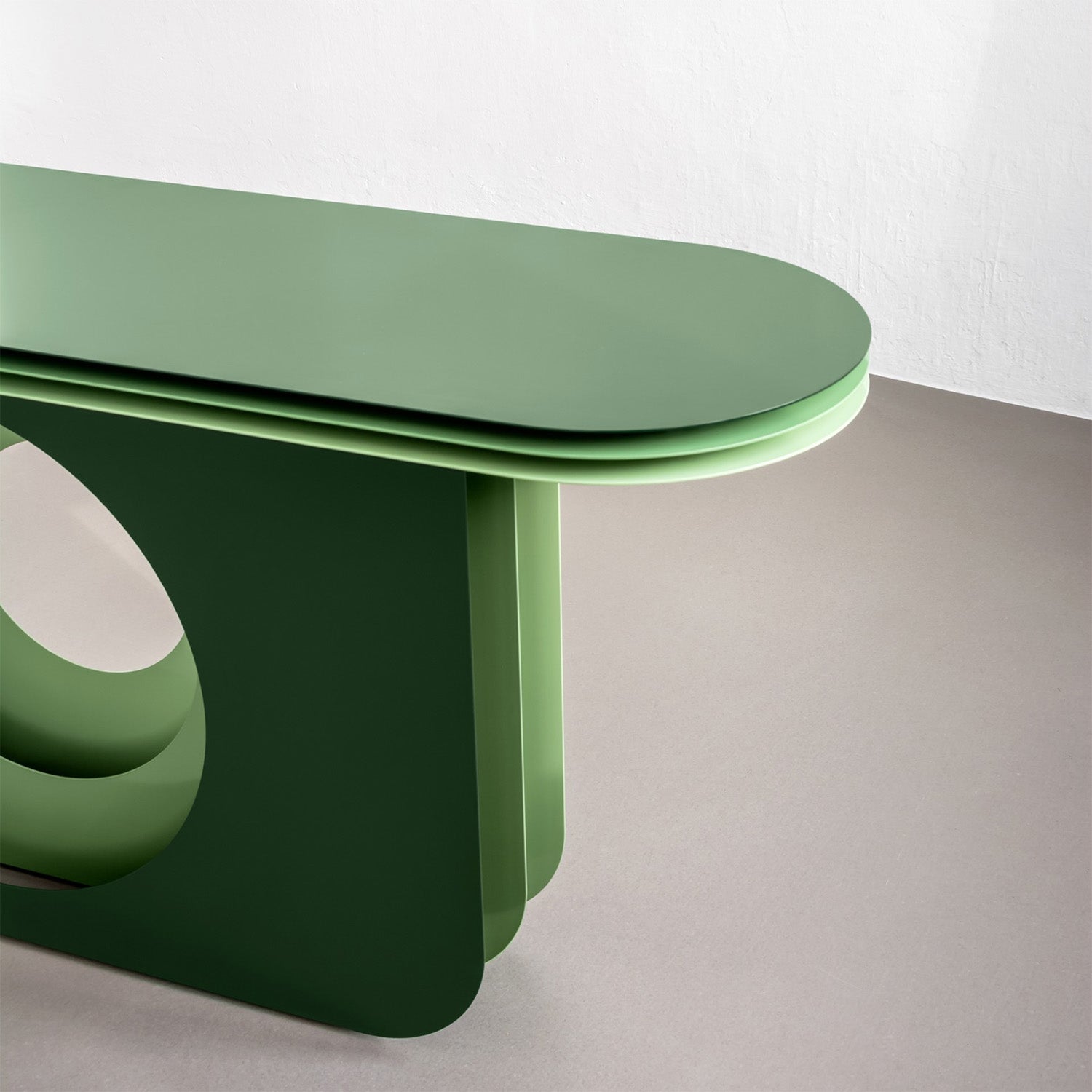 A modern, minimalist table with a sleek green finish stands against a plain white wall, offering visual depth. With rounded edges and an artistic design featuring overlapping panels and a circular cutout at the base, it evokes the elegance of Victor Foxtrots My Ami Sideboard.