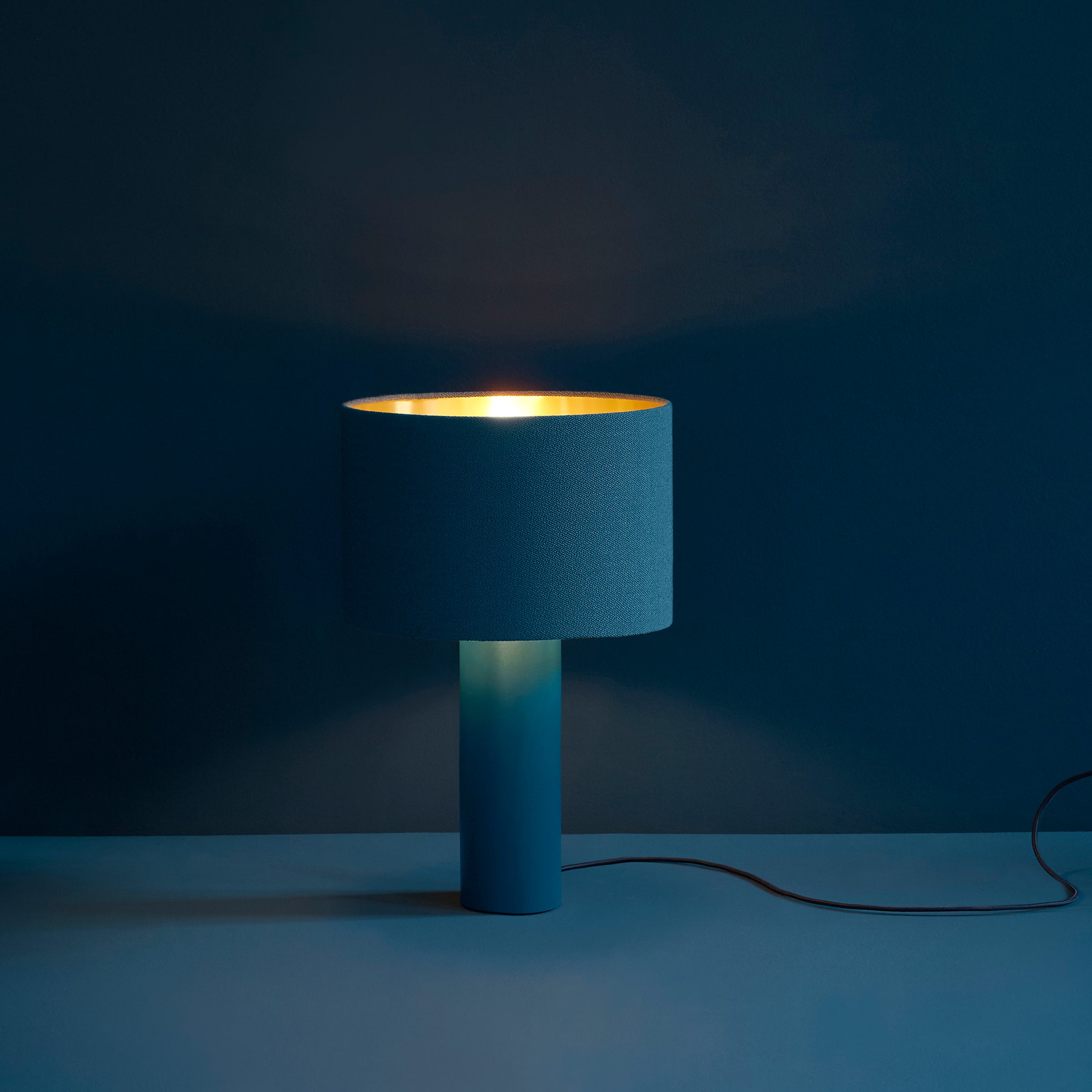 The ALL ROUND Table Lamp by Victor Foxtrot features a minimalist modern design with a cylindrical base and shade, emitting soft, warm light. Both the lamp and wool fabric background are deep blue, with a contrasting glow and an offside cord.