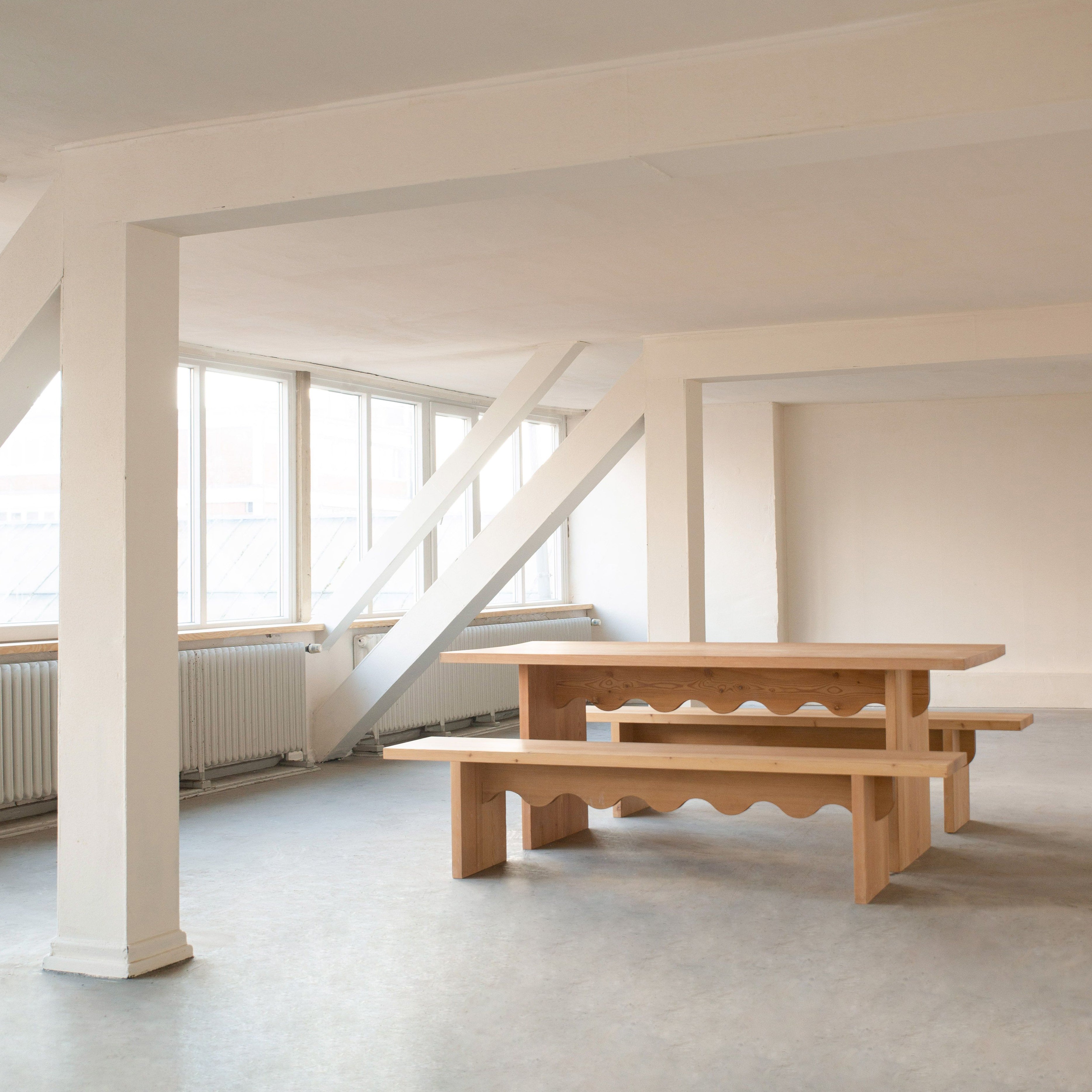 In a minimalist interior, the Schneid Tami Dining Collection stands out with its naturally oiled larch wood table and benches with wavy cutout details. Large windows, white walls, and exposed beams enhance the modern, airy ambiance.