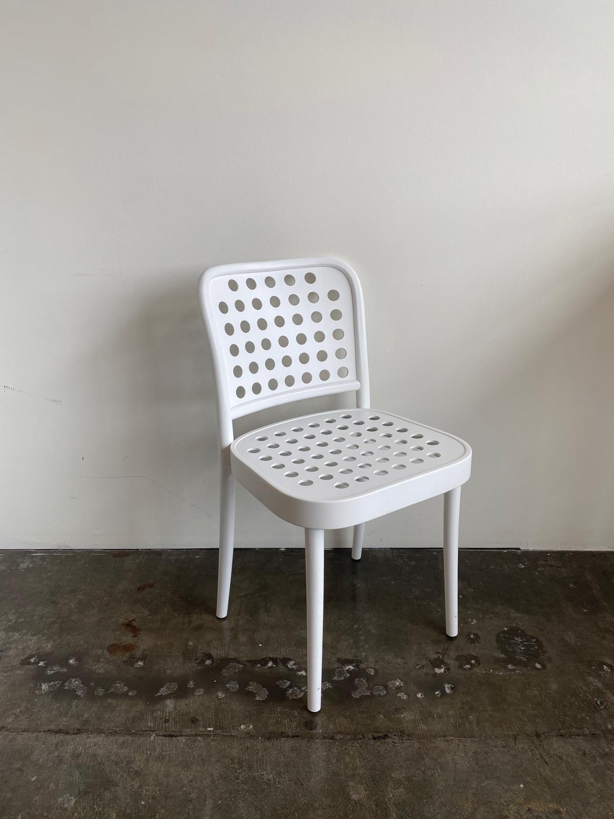 The Chair 822 by TON, a white chair with circular perforations in its back and seat, embodies Scandinavian minimalism. It elegantly stands on a concrete floor against a light wall, reflecting Claesson Koivisto Runes sleek design aesthetic.