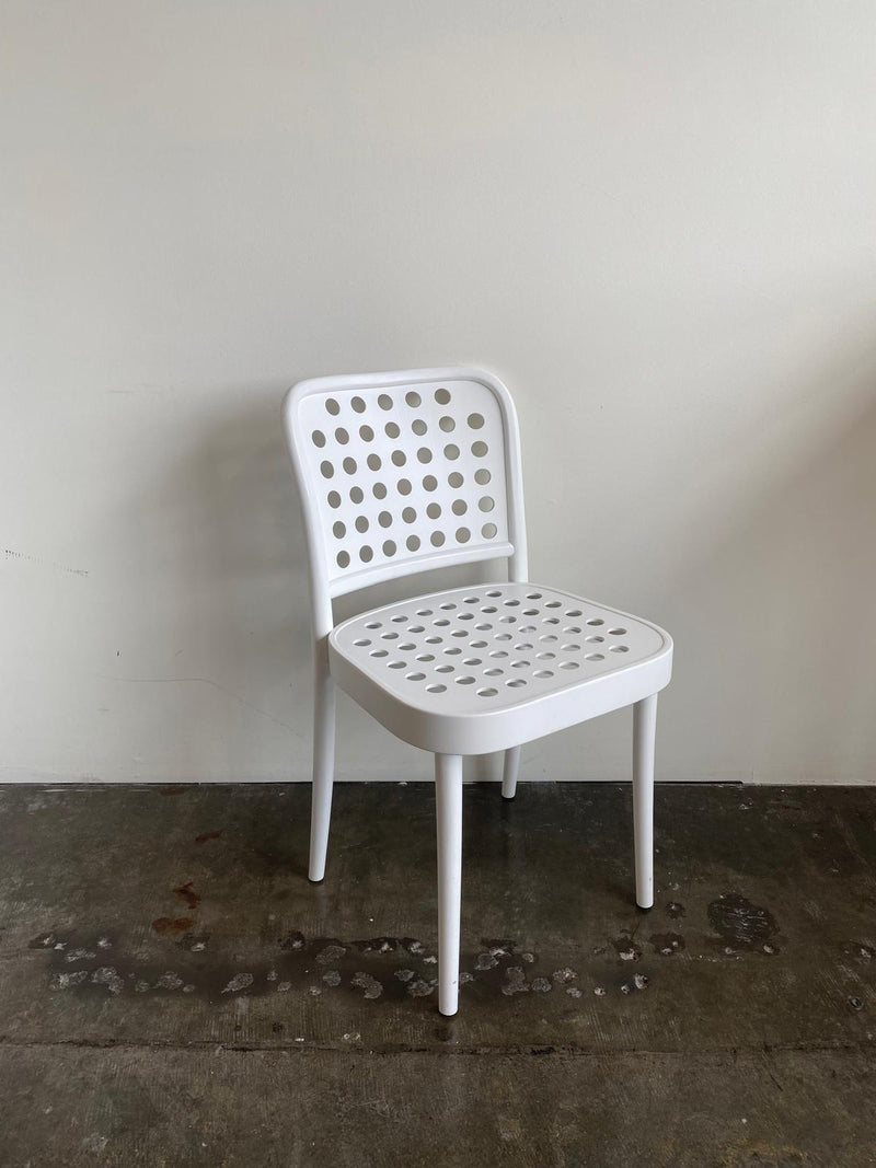 The Chair 822 by TON, a white chair with circular perforations in its back and seat, embodies Scandinavian minimalism. It elegantly stands on a concrete floor against a light wall, reflecting Claesson Koivisto Runes sleek design aesthetic.