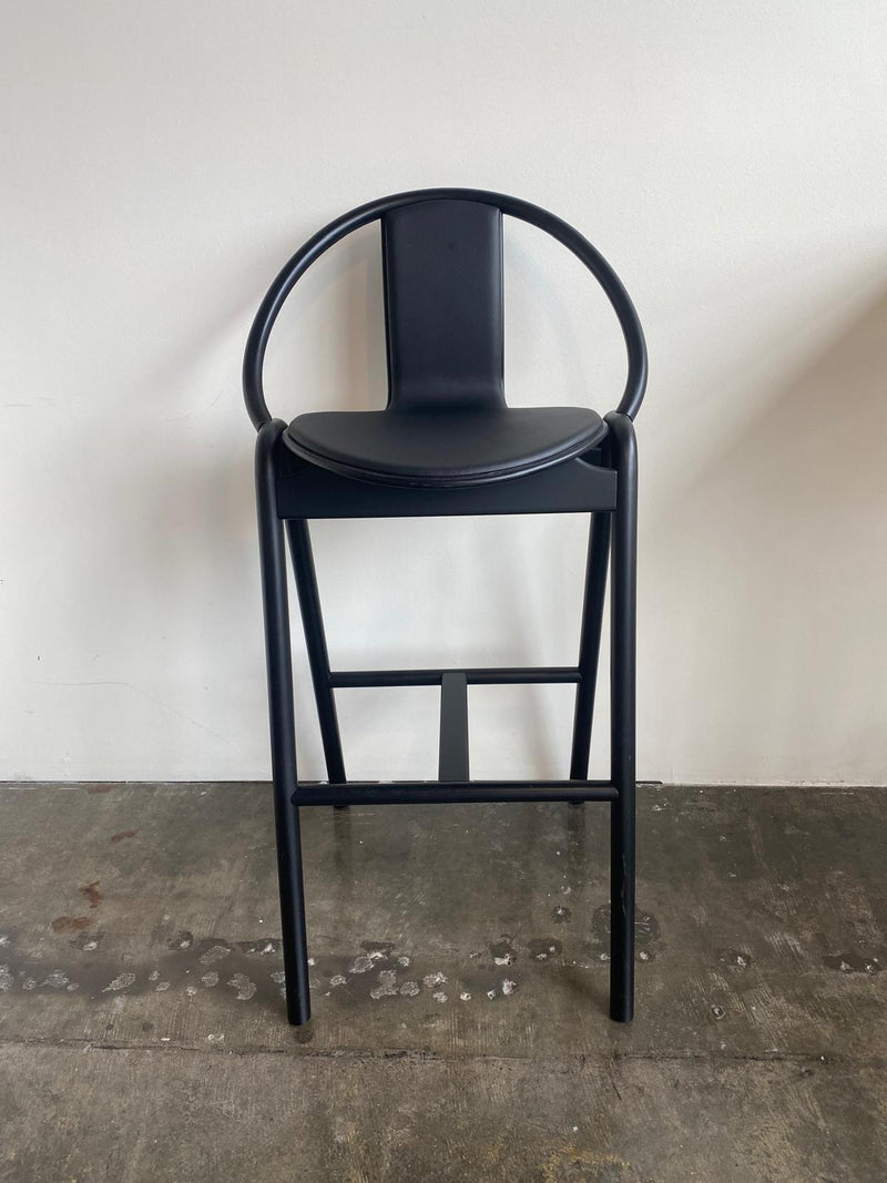 The Barstool Again by TON, with its circular backrest and minimalist design, stands on a concrete floor against a white wall. Its sleek lines, dark finish, and bentwood frame create a contemporary look.