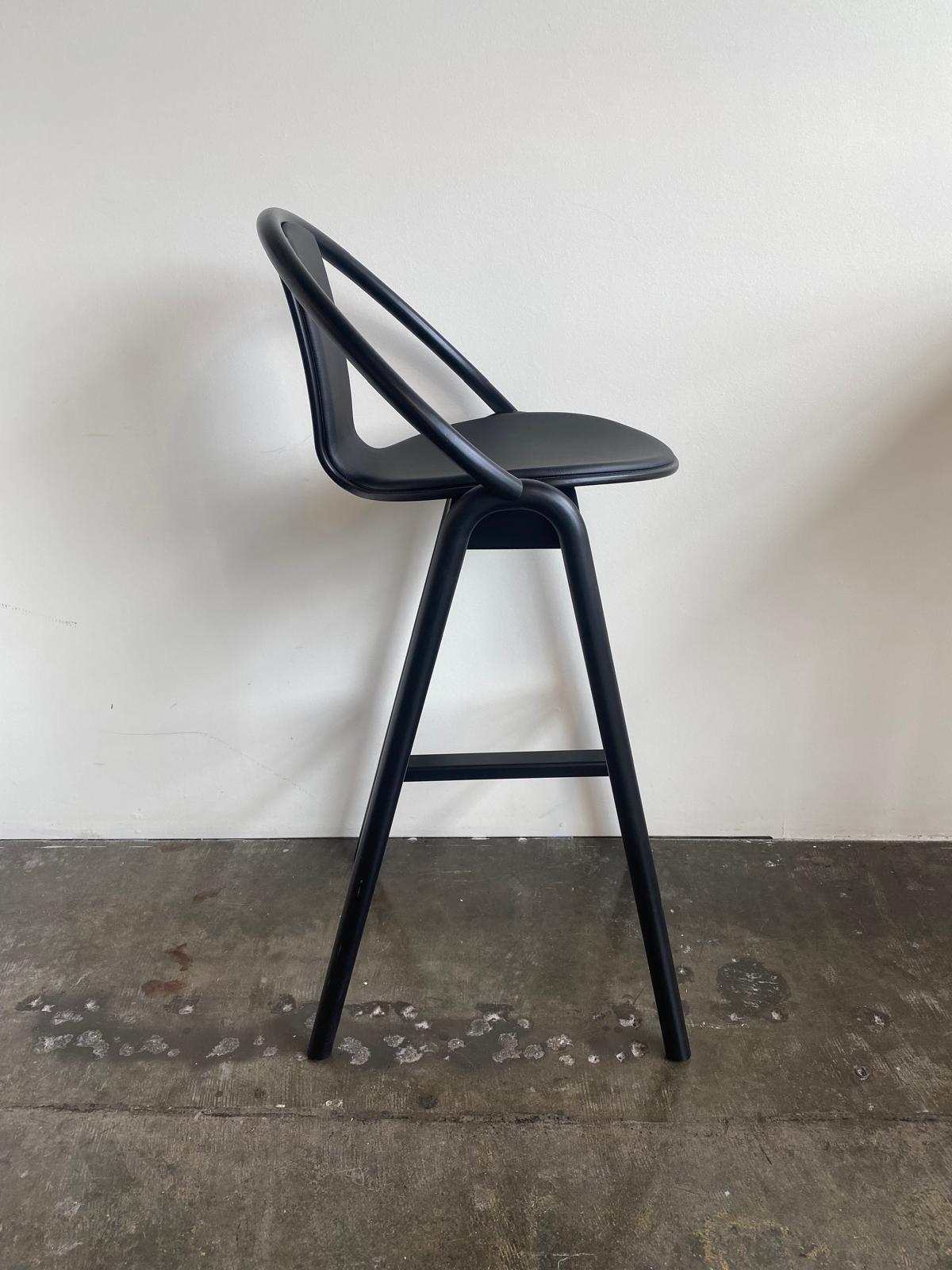 The Barstool Again by TON features a sleek, modern design with a black hue, curved bentwood backrest, and slender legs. Its elegantly positioned on a concrete floor against a plain white wall.