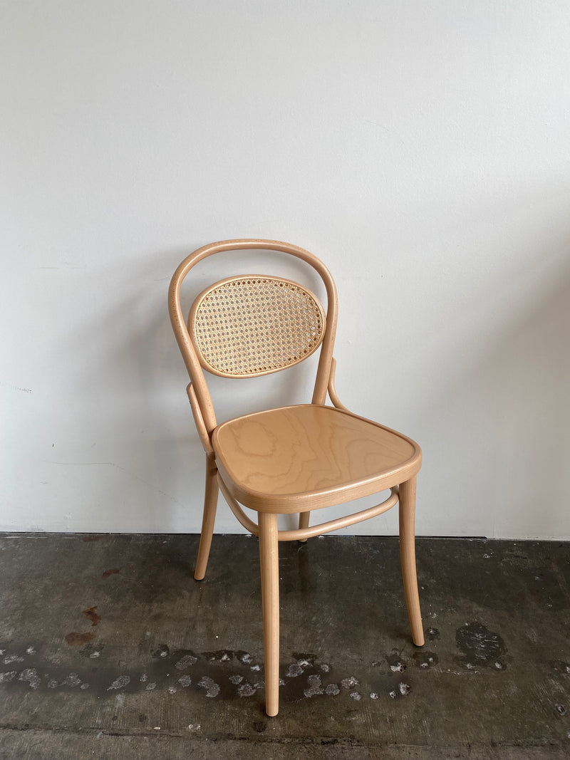 In front of a plain white wall, the Chair 20 Cane-Sale by TON sits elegantly on the concrete floor. Its light beech frame and graceful curved design enhance the minimalist space with sophistication.