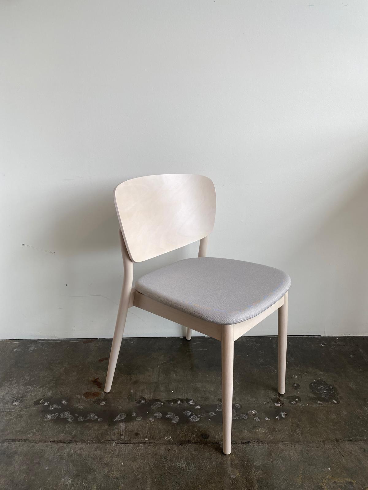 The Chair Valencia Upholstered by TON is a light wooden chair with an ergonomic design, featuring a wide curved backrest and a light gray upholstered seat, placed on a concrete floor against a white wall.