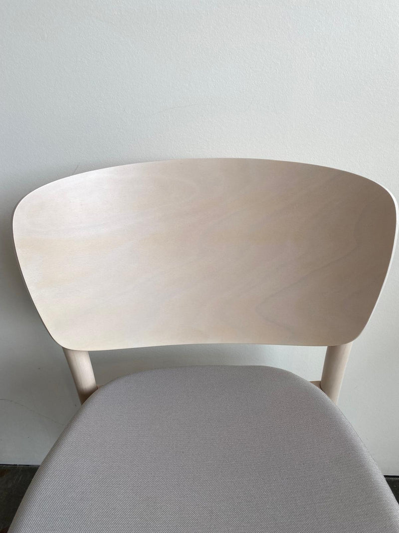 The Chair Valencia Upholstered by TON is a minimalist wooden chair with solid construction, featuring a smooth, light-colored backrest and cushioned seat. This timeless piece is ideal against a plain, light-colored wall.