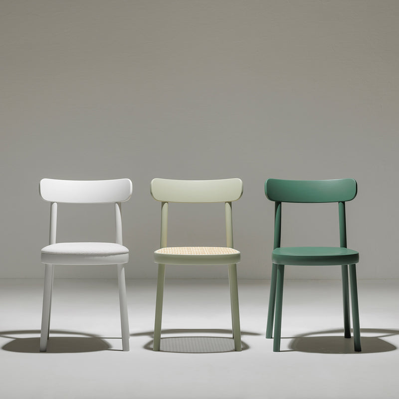 In a minimalist setting, the Chair La Zitta by TON showcases three designs: a white chair with a cushioned seat, a light green chair with a woven seat, and a dark green chair with solid wood. Each features simple curved backrests reflecting Alex Guflers ergonomic design principles in the well-lit, neutral space.