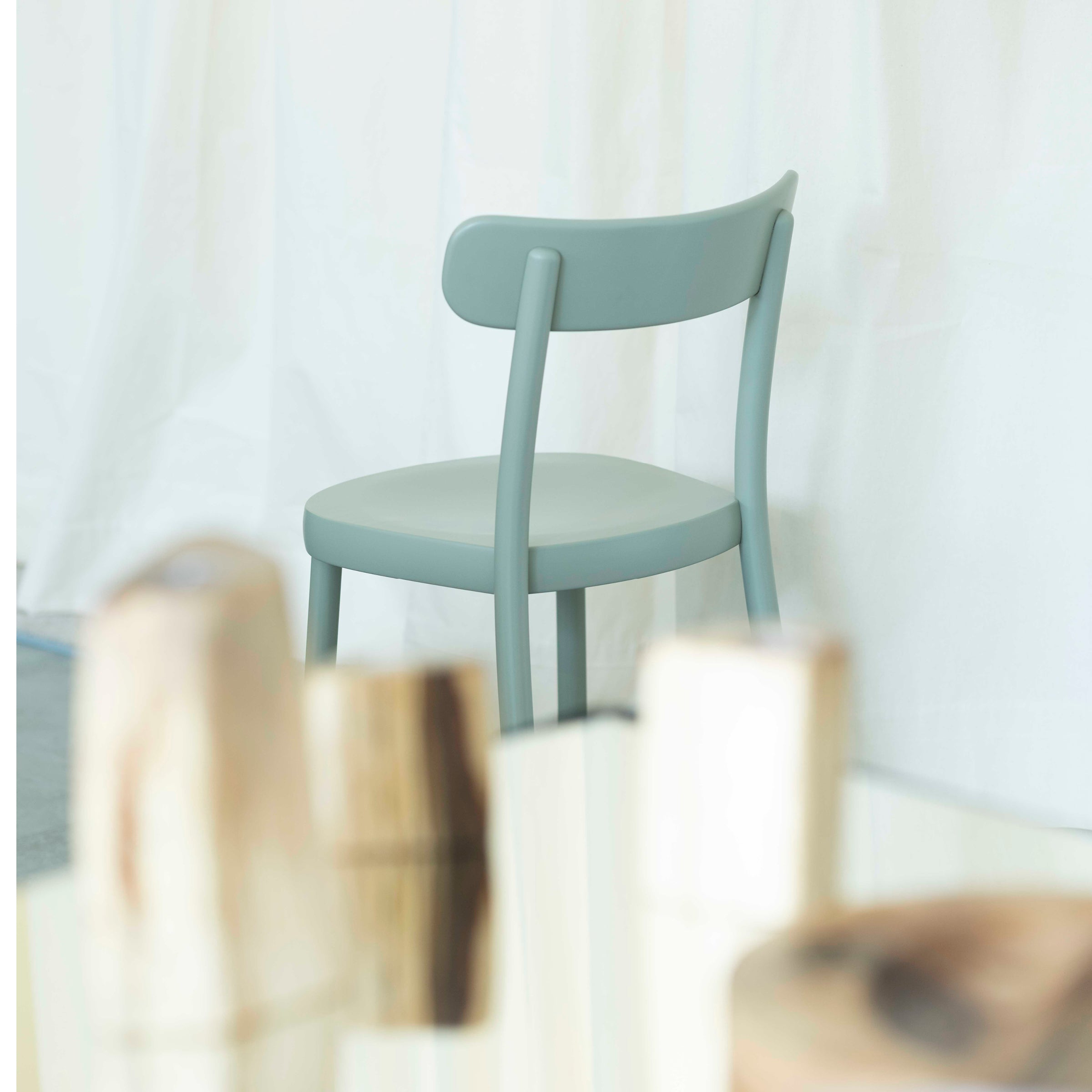 On a light floor, partially obscured by blurry cylindrical objects in the foreground, sits the Chair La Zitta by Alex Gufler for TON. It features a light green shade, an ergonomic design with a curved backrest, and is set against a soft white curtain background.