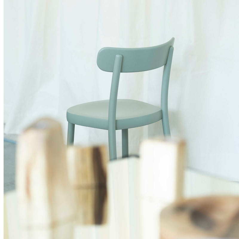 On a light floor, partially obscured by blurry cylindrical objects in the foreground, sits the Chair La Zitta by Alex Gufler for TON. It features a light green shade, an ergonomic design with a curved backrest, and is set against a soft white curtain background.