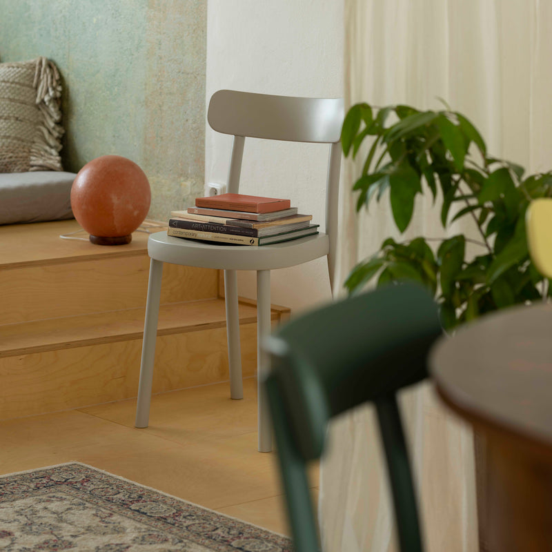 In a minimalist room, the Chair La Zitta by TON in light gray with an ergonomic design holds a stack of books by wooden steps. A large orange sphere decor is beside it, along with a green plant and part of an Alex Gufler dining set in solid wood construction visible in the foreground.