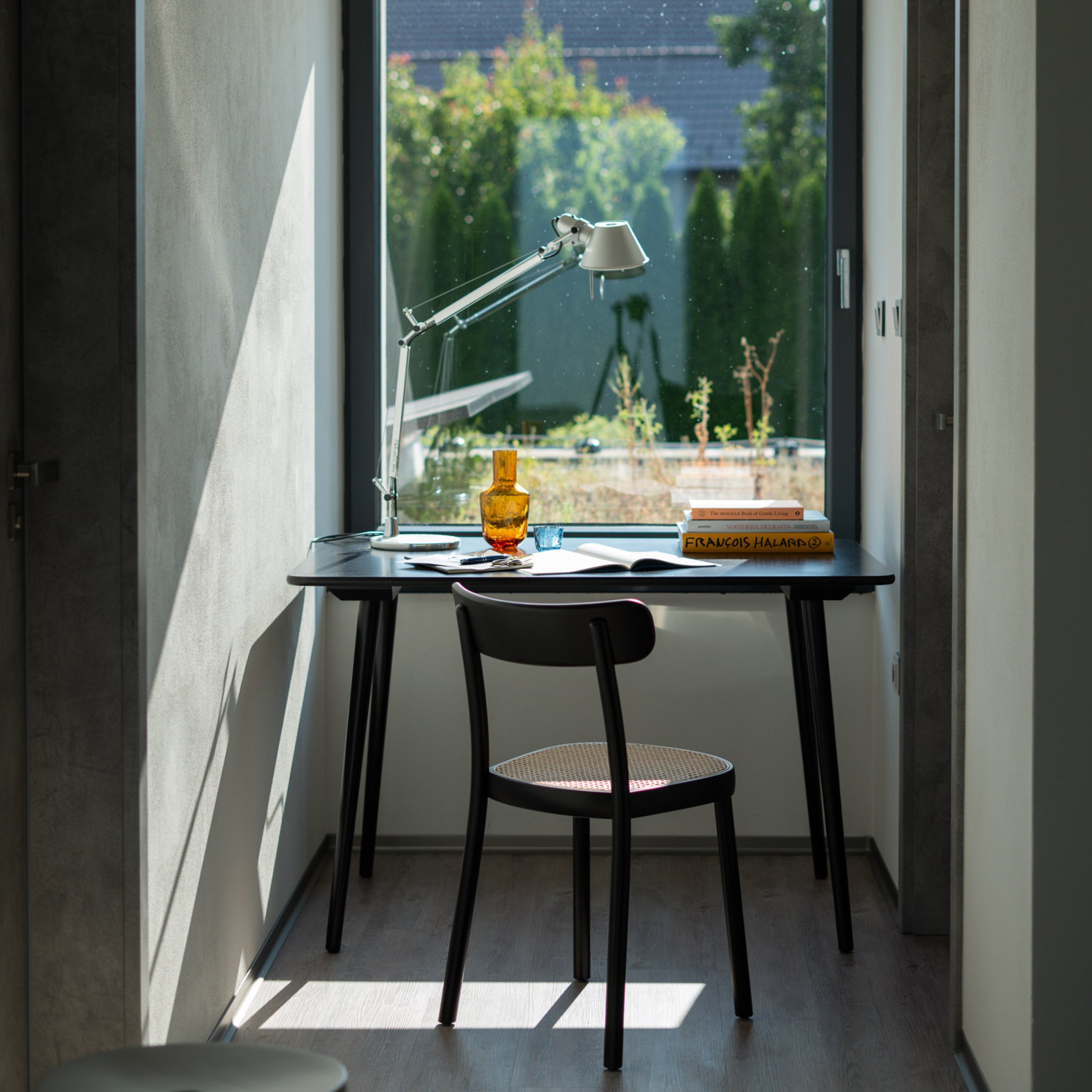 By a large window, a workspace contains a black desk and the ergonomic Chair La Zitta - Cane by TON. A white lamp, an orange glass, books, and papers sit on the desk. Outside, greenery and a house can be seen under the clear sky.