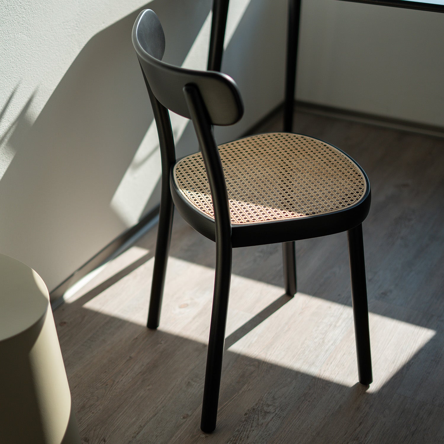 The Chair La Zitta - Cane by TON, with its ergonomic design, woven rattan seat, sleek black frame, and curved backrest, sits in a minimalist room. Bathed in sunlight through a nearby window and standing on a wooden floor, it adds to the serene ambiance.