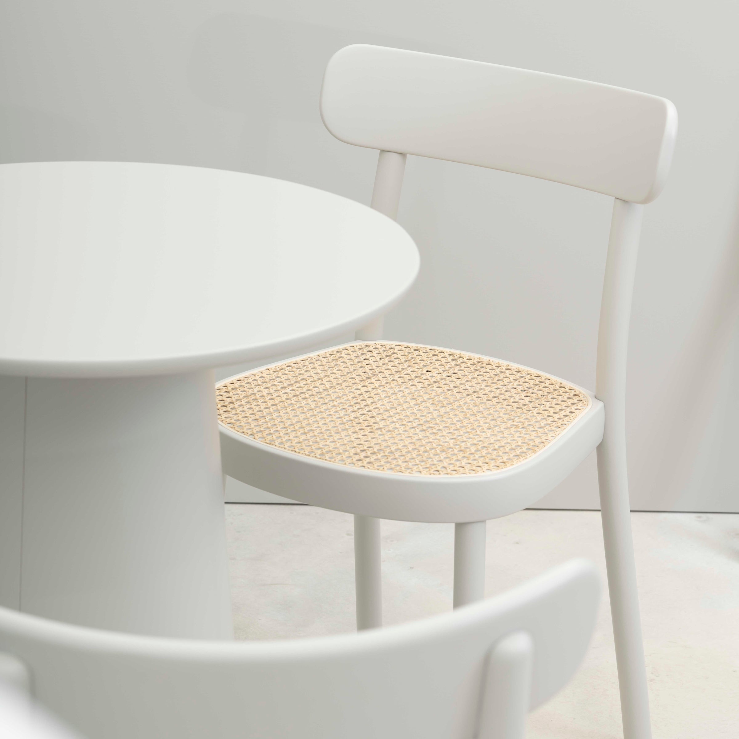 The minimalist dining area showcases a round white table paired with the ergonomically designed Chair La Zitta - Cane by TON, featuring a woven seat. The setting exudes simplicity and modernity, focusing on clean lines and neutral tones.