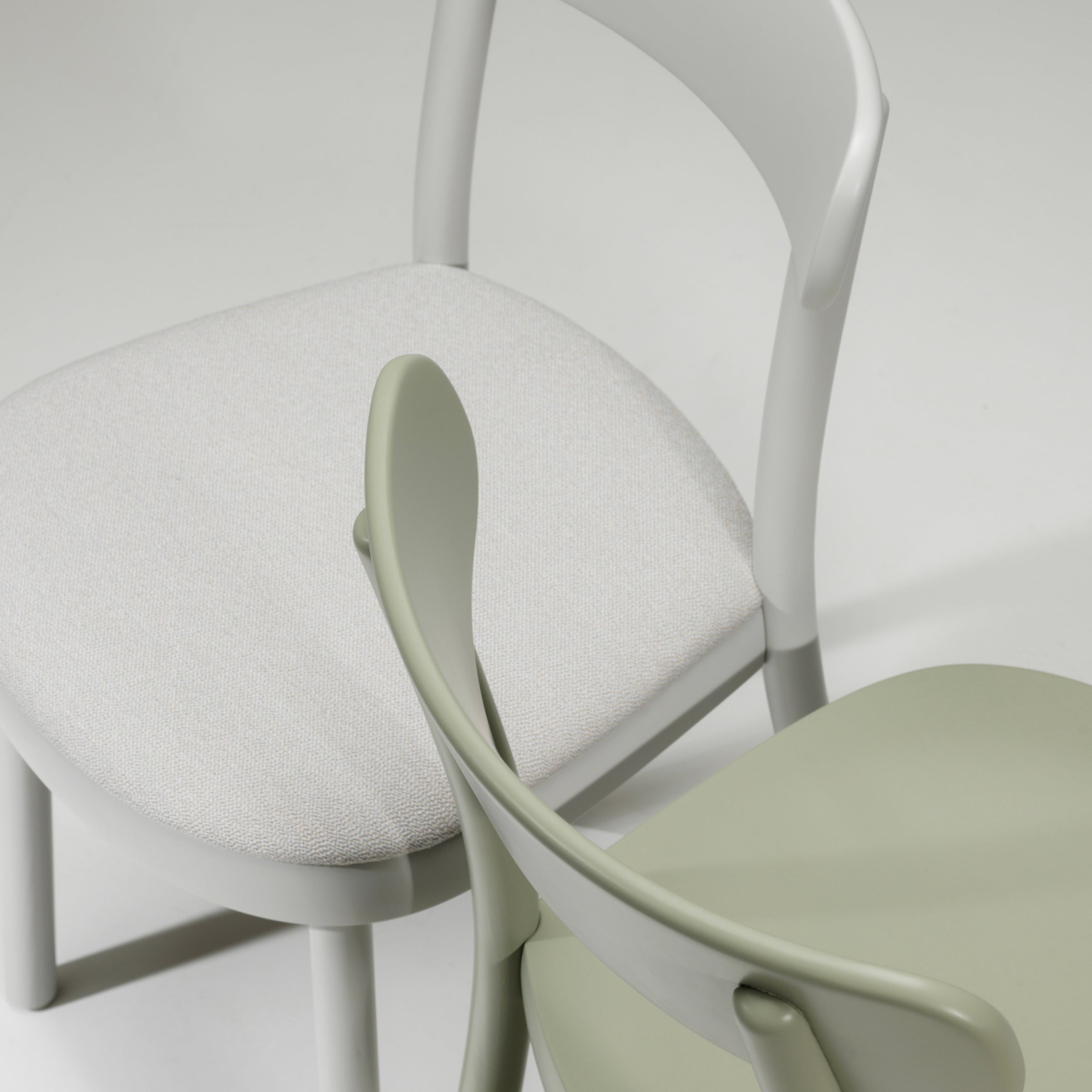 The Chair La Zitta by TON features a white fabric-upholstered seat and another one in light green with a smooth finish. Both chairs have curved backs, ergonomic designs, and solid wood construction for lasting support.