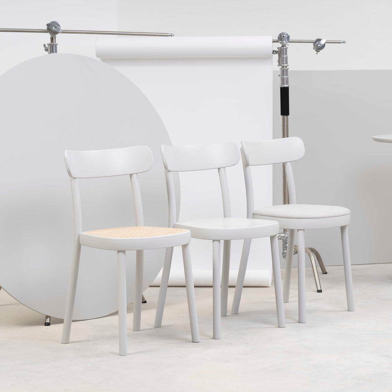 Three Chair La Zitta - Upholstered by TON, in white, are aligned against a minimalistic backdrop with a circular panel. They feature solid wood construction: two have cushioned fabric seats and one boasts an ergonomic woven texture.