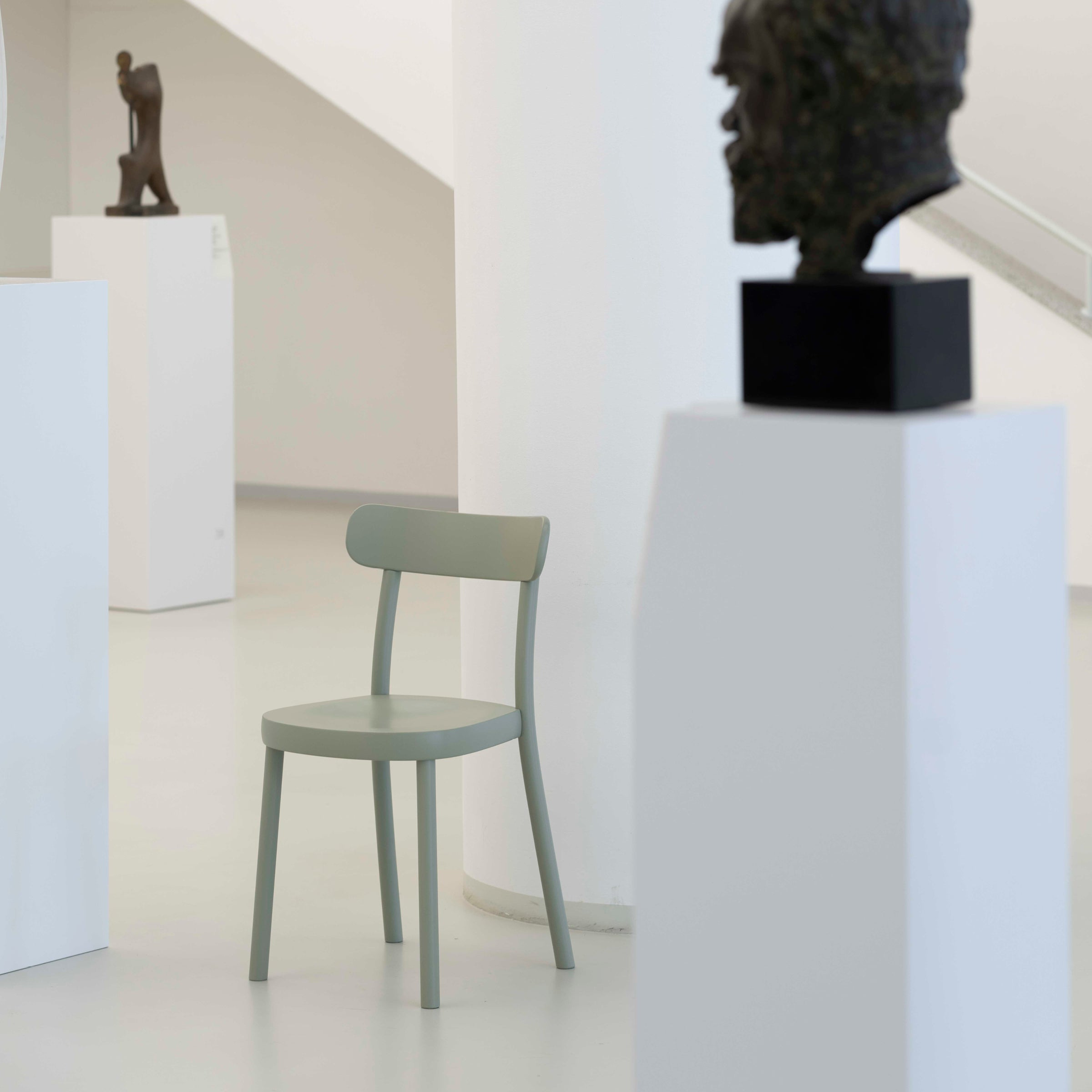 In a minimalistic museum interior, the ergonomic Chair La Zitta by TON, designed by Alex Gufler, is centrally placed among white pedestals. A bronze bust sculpture adorns the foreground, while a figurative sculpture stands in the background against immaculate white walls and floors.